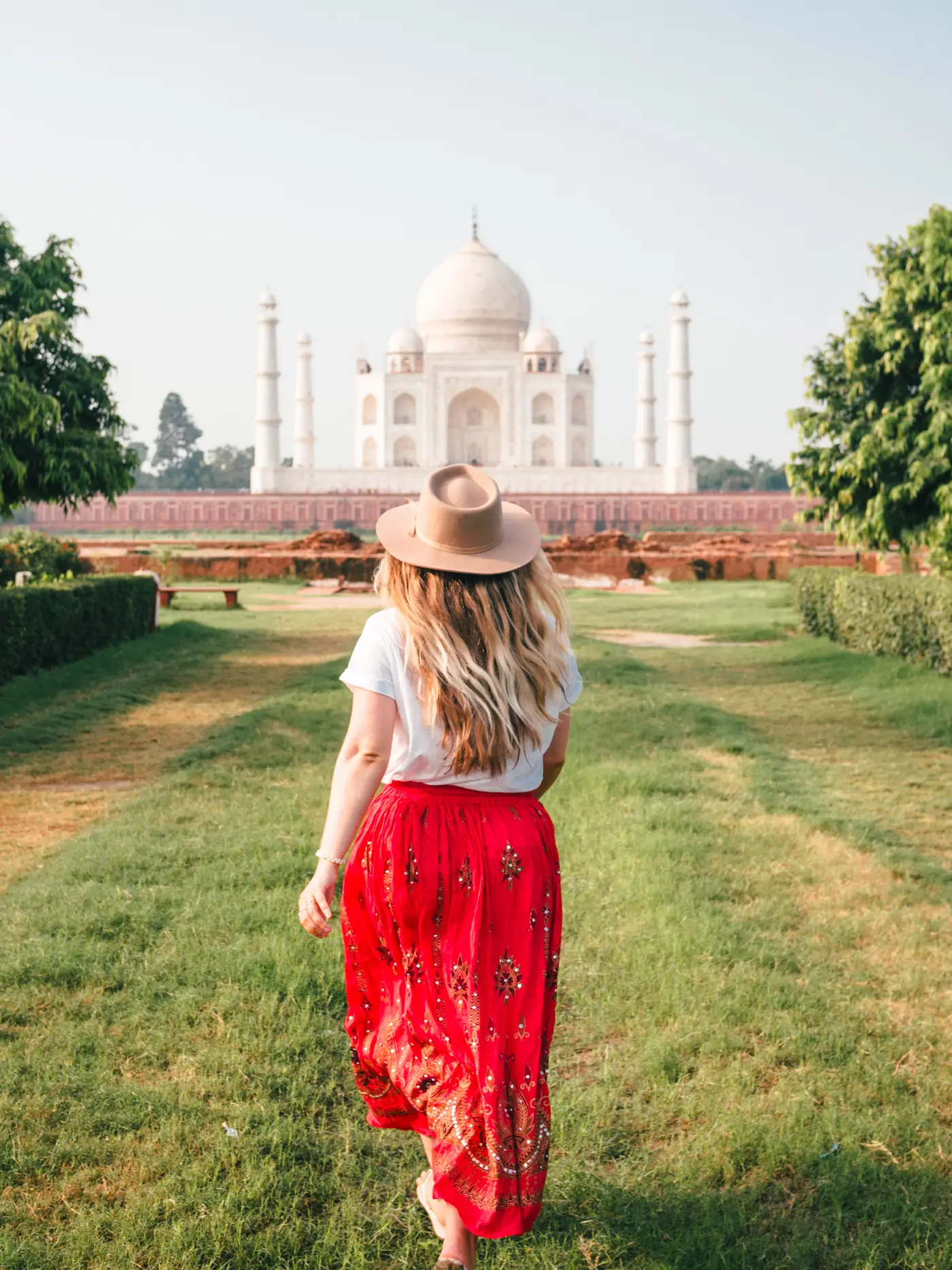 Girl with long dark blonde hair, wearing a red skirt, white t-shirt and beige hat, running towards Taj Mahal in Mehtab Bagh garden.