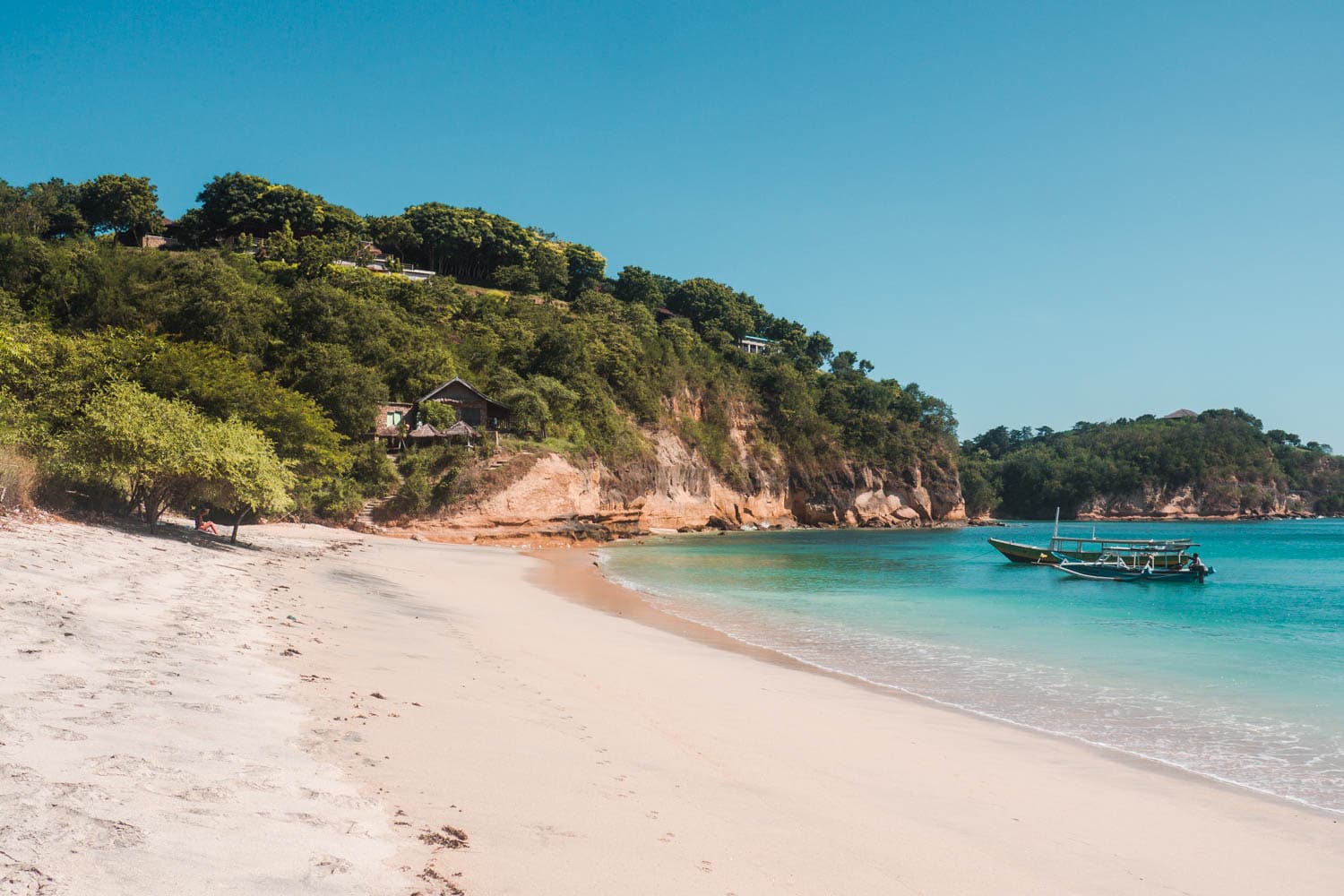 White sand beach, Pantai Rungkang, with a lush green cliff in the background and turquoise water and blue skies, one of the top things to do and best beaches in Lombok. 