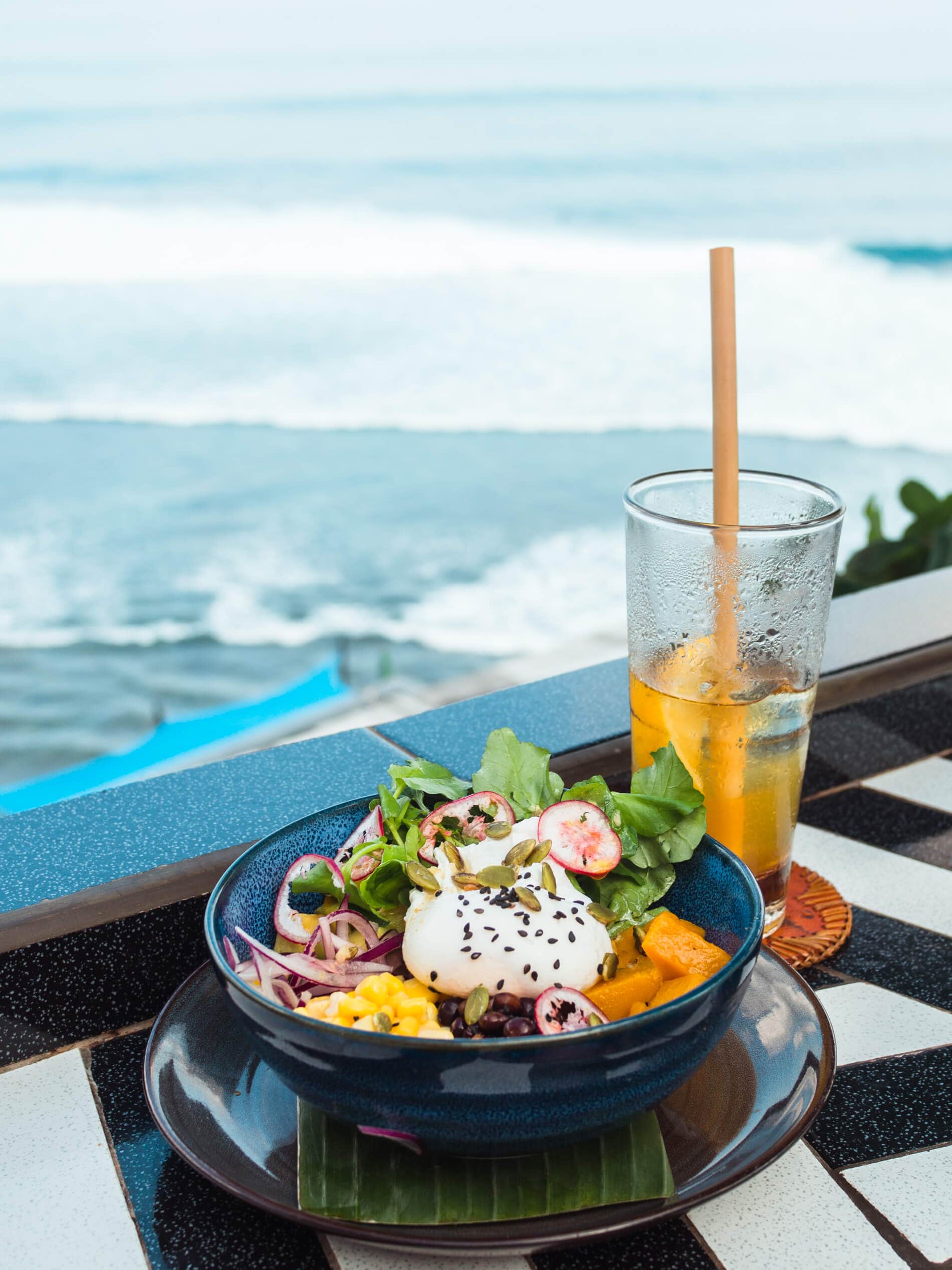 Black bowl of breakfast with egg and vegetabkes with an iced tea with Uluwatu waves in the background, one of the top things to do in Bali.