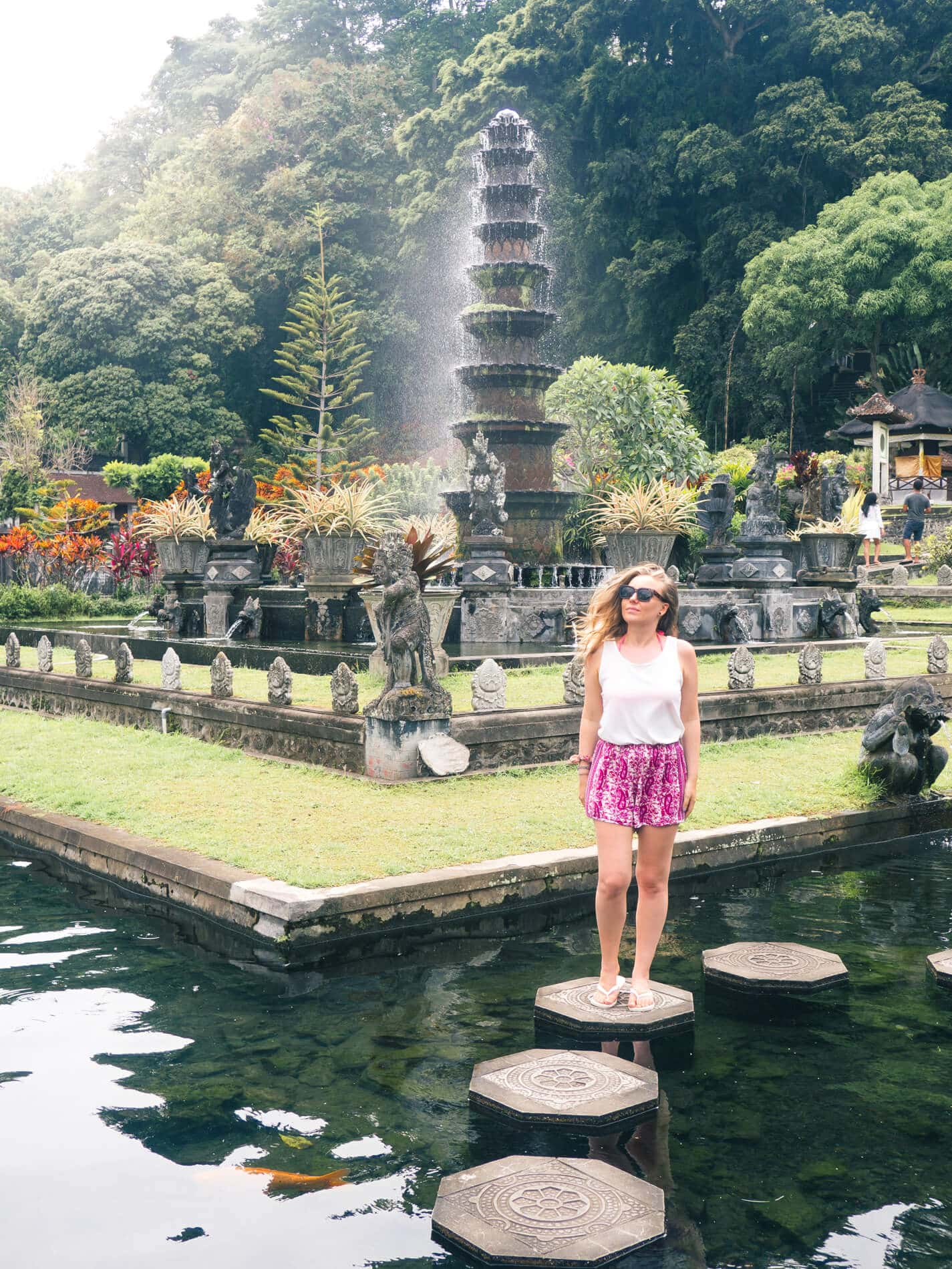 Woman, wearing a white top and purple shorts, standing on a stone in one of the Koi ponds at Tirta Gangga Water Palace, a must on any Bali Bucket List.