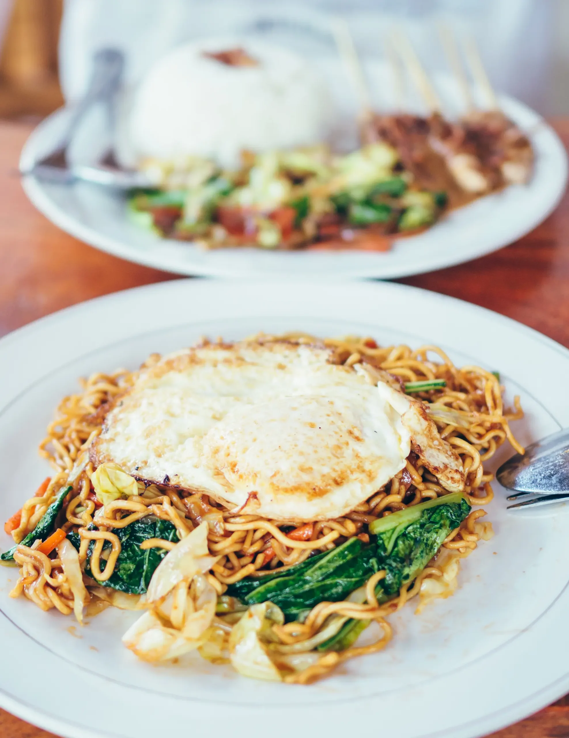 White plate with noodles Mie Goreng with a fried egg on top, white plate with rice and chicken sate in the background, on the the top things to do in Bali.