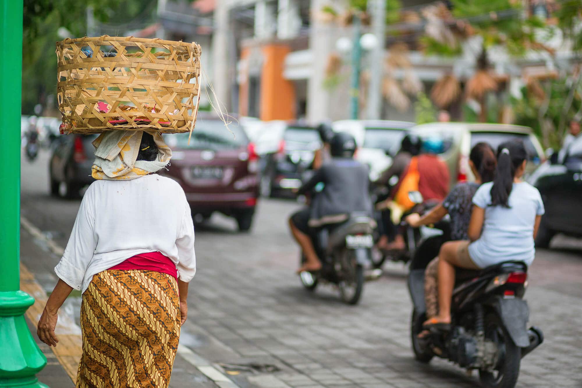 A busy street in Ubud with cars, motorbikes and a Balinese woman walking with a basket on her head. How to get around Ubud during this Ubud itinerary
