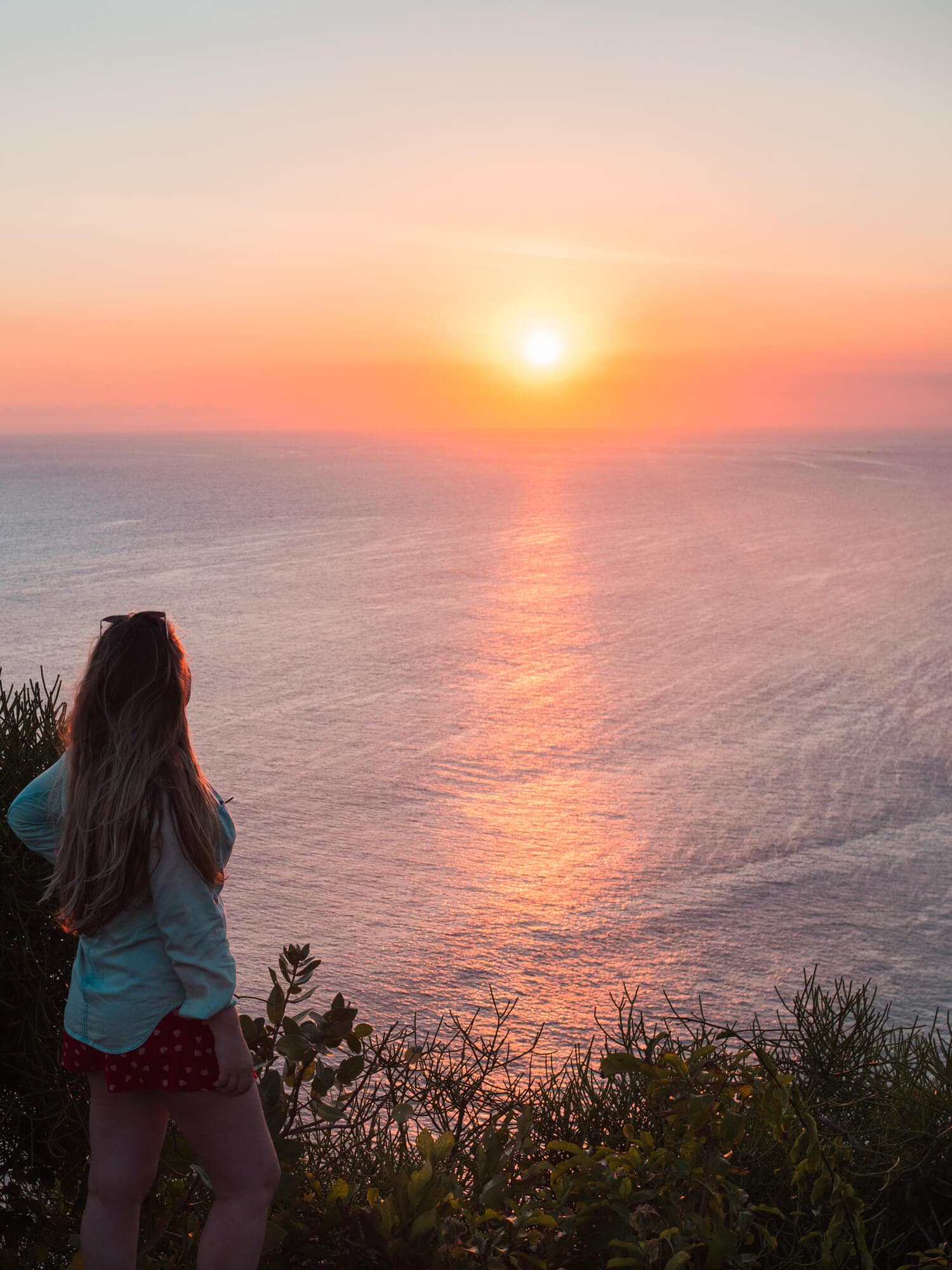 Girl watching the sunset from Karang Boma Cliff in Uluwatu. The sky is pink and orange and the sun bright yellow.