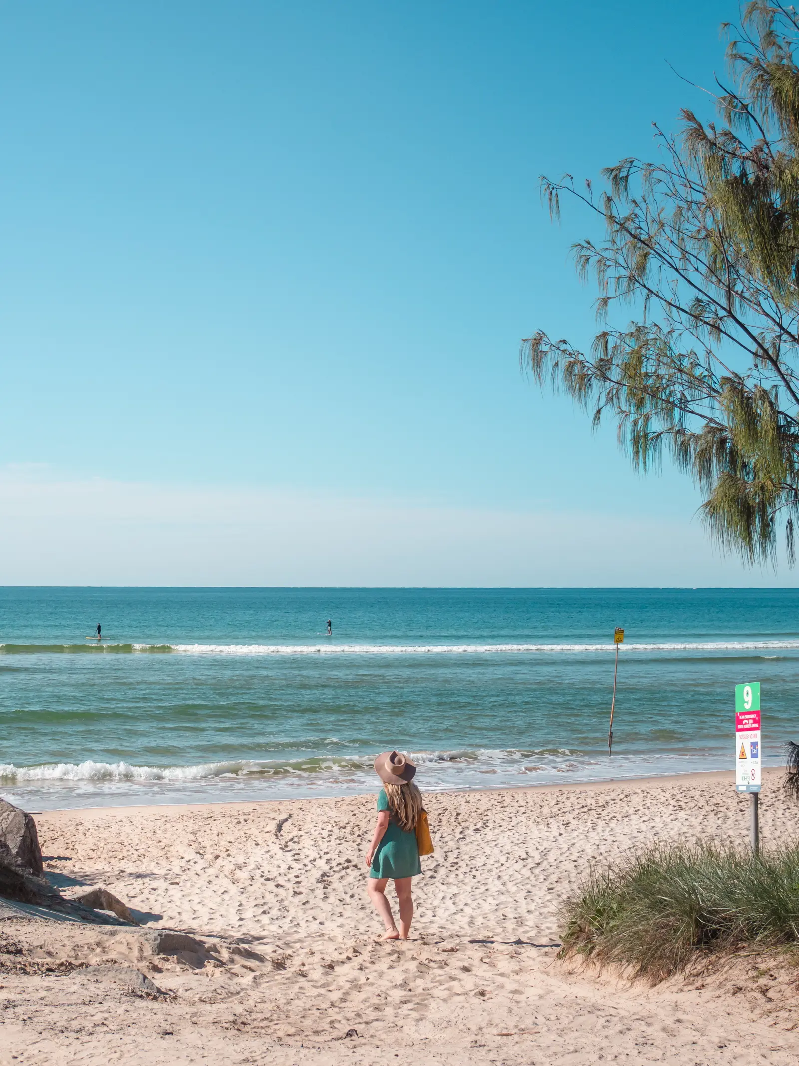 Girl in a green dress and beige hat standing on Noosa Main Beach looking out at the ocean.