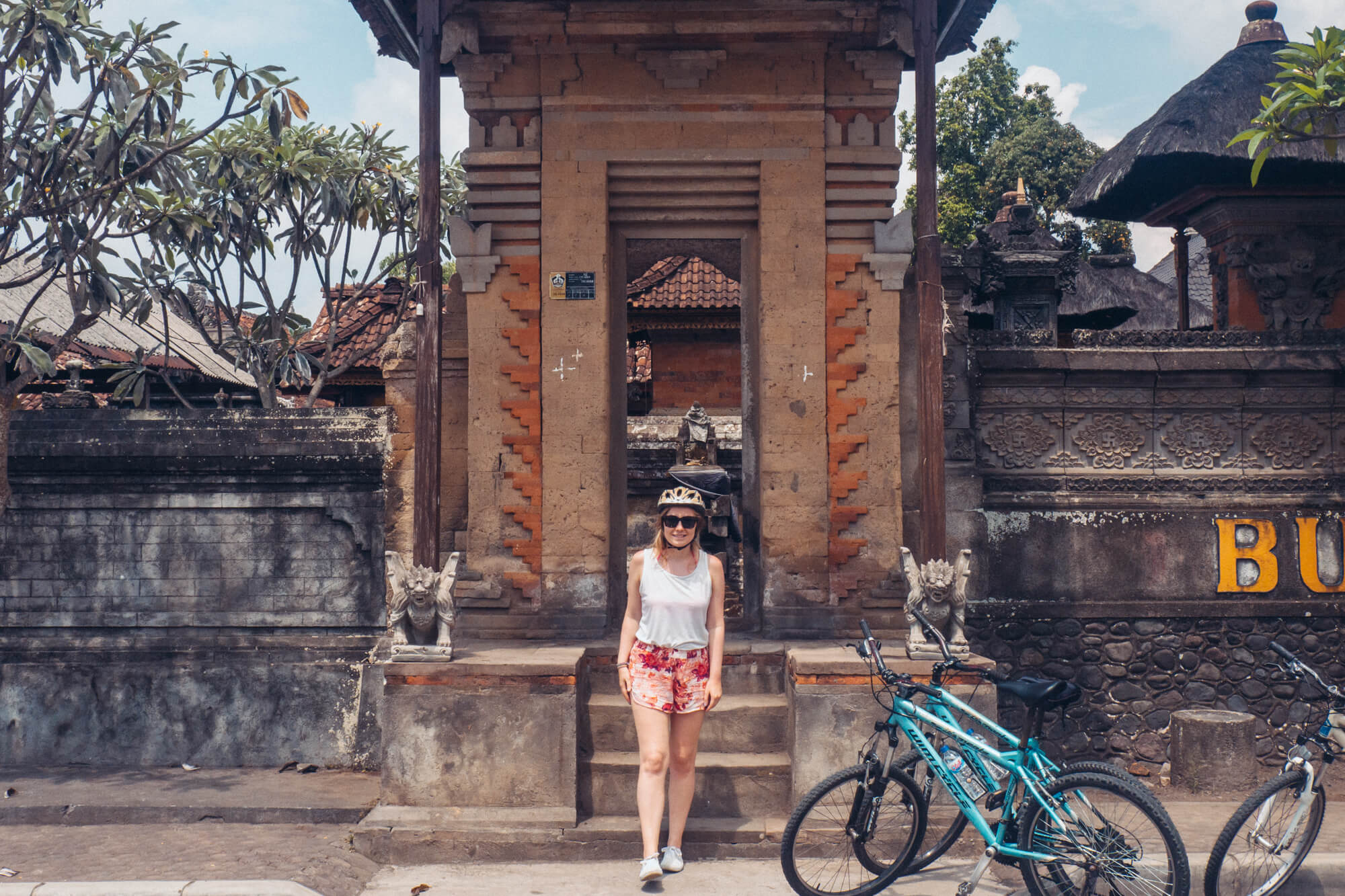 Girl in pink shorts and a white top standing in front of a light brown temple entrance during a cycling tour in Bali.