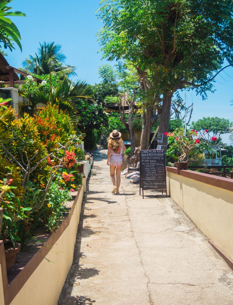 Girl in a hat and tie dye playsuit walking along the Jungut Batu Boardwalk, lined by orange flowers and greenery, on a day trip to Nusa Lembongan.