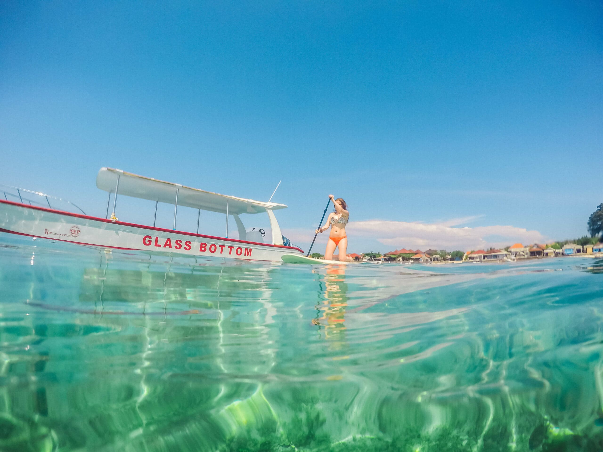 Girl paddle boarding on clear turquoise water with a glass bottom boat in the background on a day trip to Nusa Lembongan.