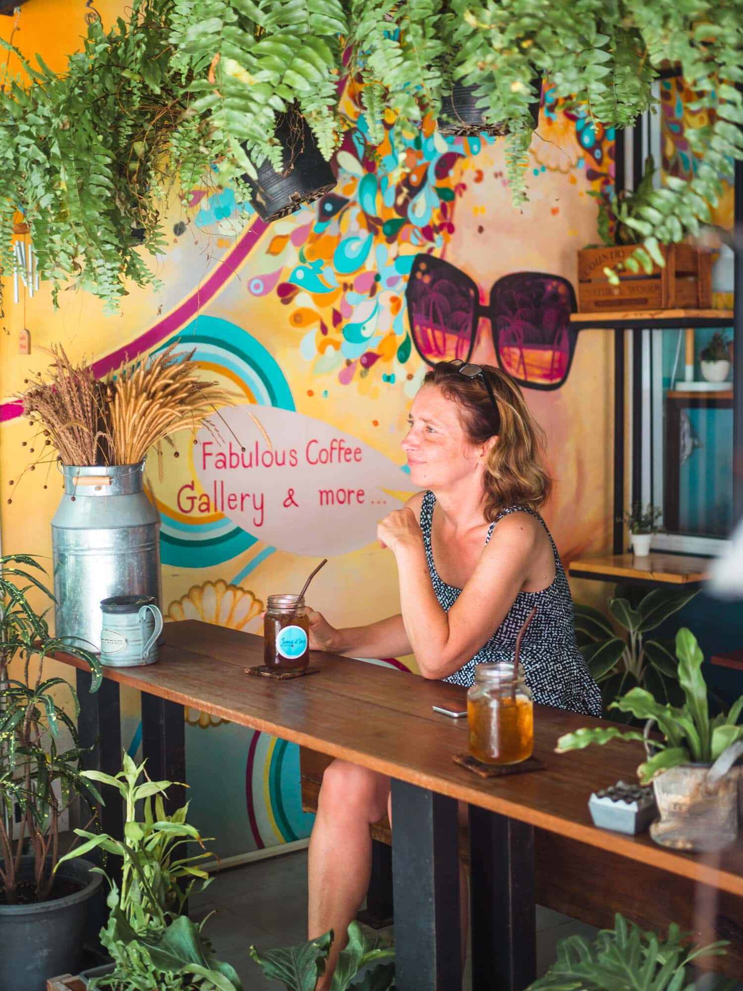 Woman sitting at a wooden table with an iced tea in hand and a yellow mural background at Jump at Seas café on Koh Samet.