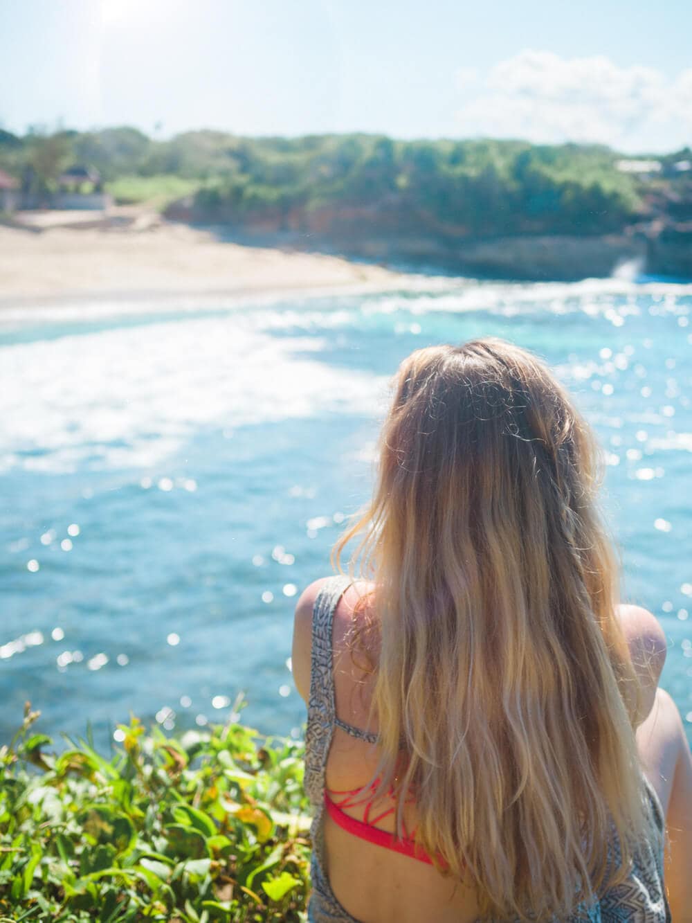 Girl with long hair wearing a red bikini and grey dress looking out over the turquoise water towards Dream Beach on a day trip to Nusa Lembongan.