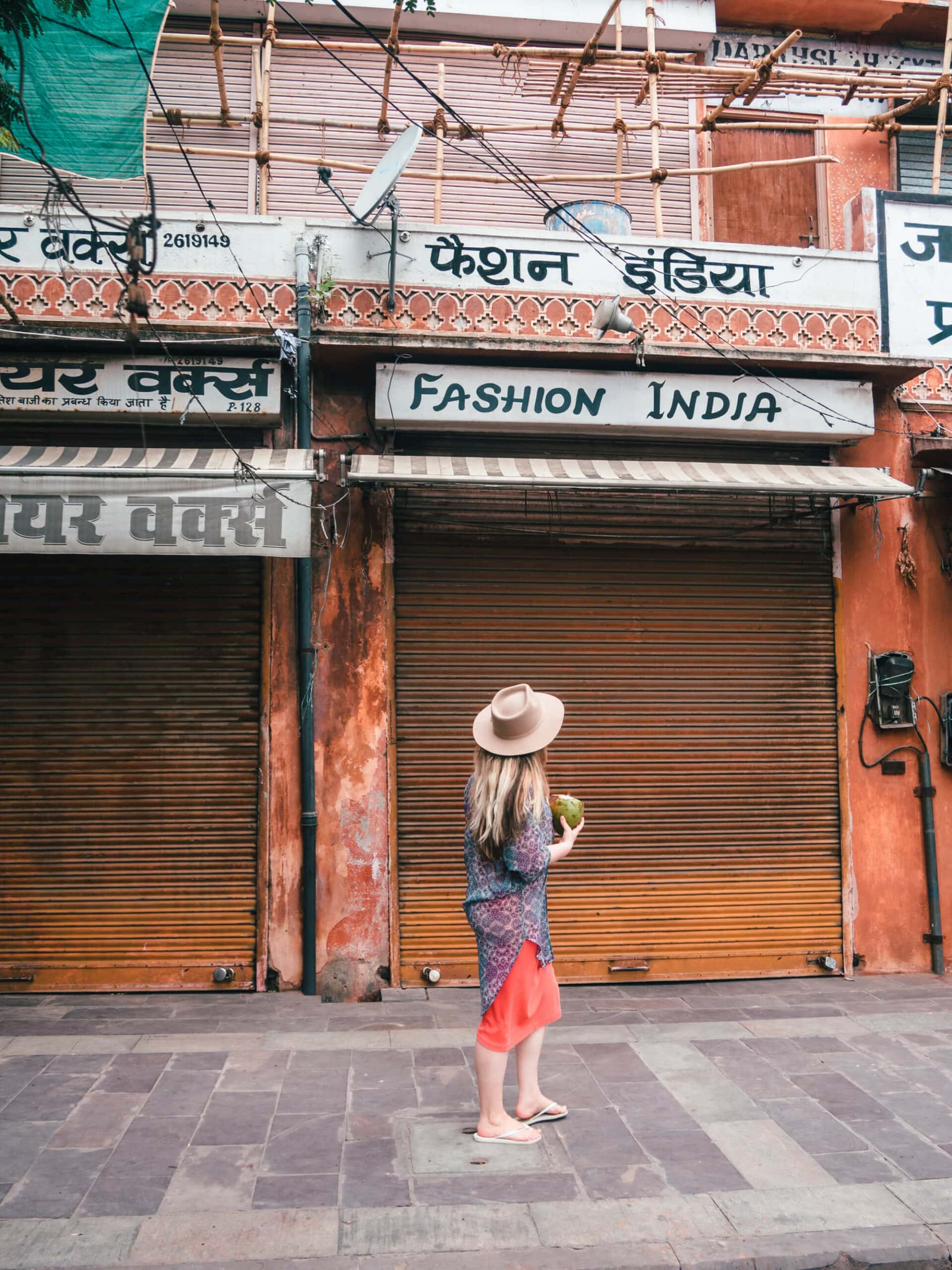 Girl in a coral dress and blue kimono wearing a beige hat and holding a coconut exploring the streets on Jaipur, a must on any 2-day Jaipur itinerary.