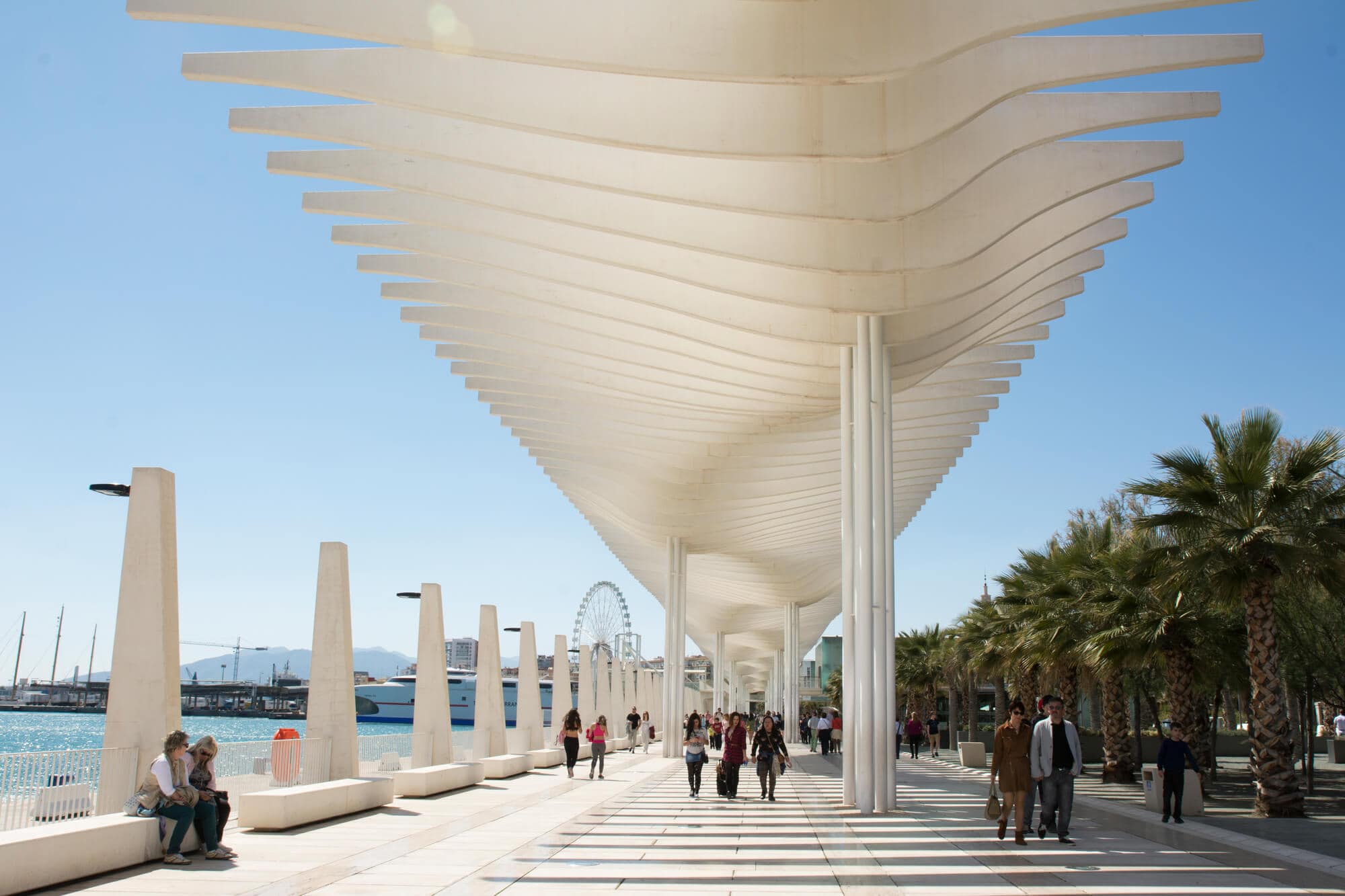 People walking along the Muelle Uno area of Malaga Cruise Port on a sunny day, under a white sun shading architectural sculpture.