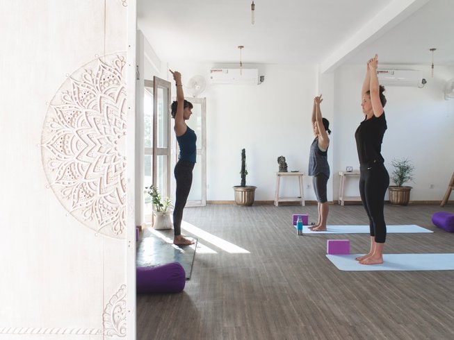 Three women in dark yoga attire standing on turquoise yoga mats in a white room with wooden floor, and a white door with Balinese carvings, during a surf and yoga retreat in Bali.