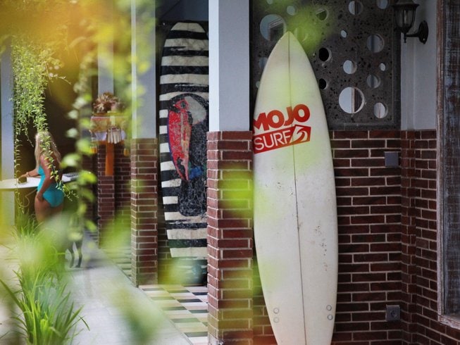 White surfboard with Mojosurf camp logo standing against a brick wall, blurry  green and yellow leaves in the foreground, on a surf and yoga retreat in Bali.
