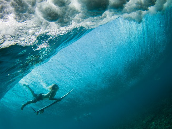 Boy in boardshorts with a small surfboard duck diving under a large blue wave during a surf and yoga retreat in Bali.