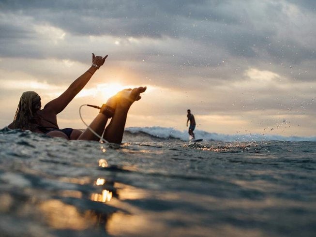 Woman on a surfboard with one hand in the air in the water during sunset, in the background is a man surfing a wave, during a surf and yoga retreat in Bali.