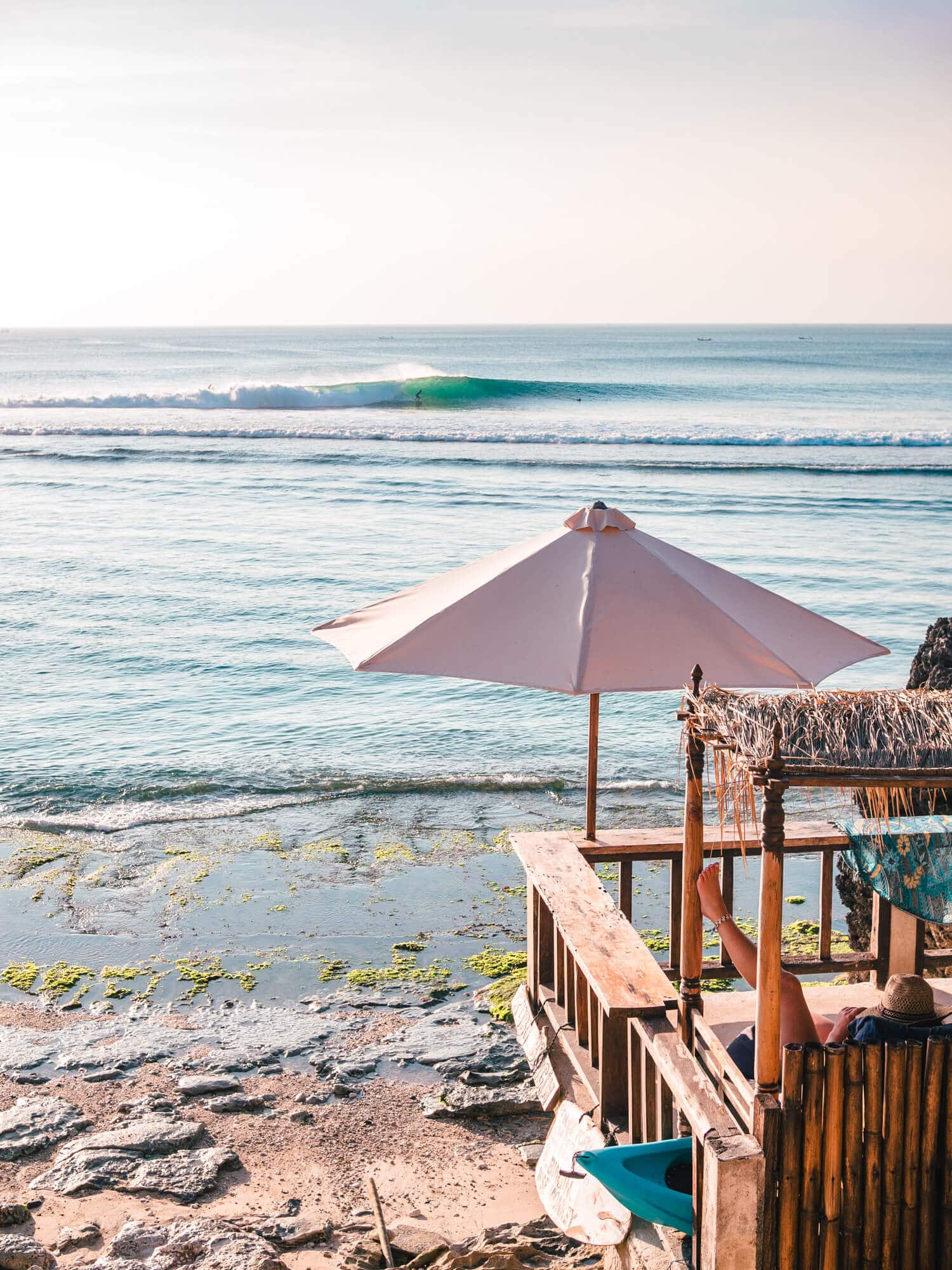 Woman sitting on a wooden veranda under a white parasol looking towards a surfer on a wave early in the morning at Bingin Beach, one of the best places for a surf and yoga retreat in Bali.