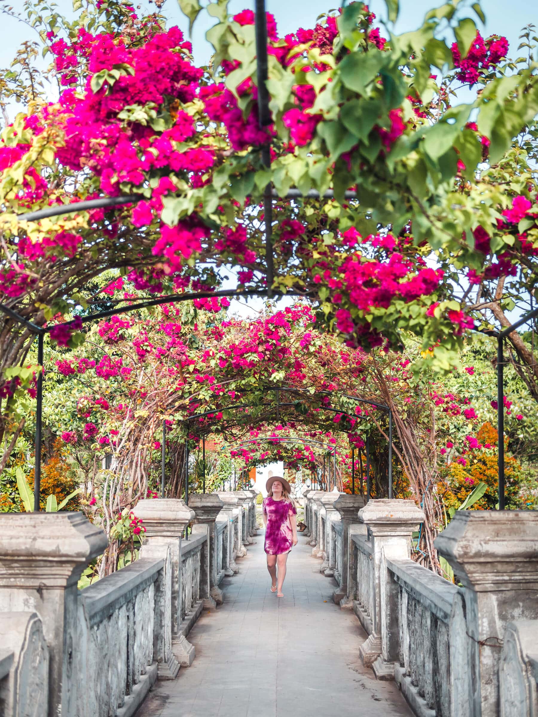 Girl in a purple dress and beige hat walking over a bridge in Taman Ujung Water Palace in East Bali during a two week Bali itinerary.