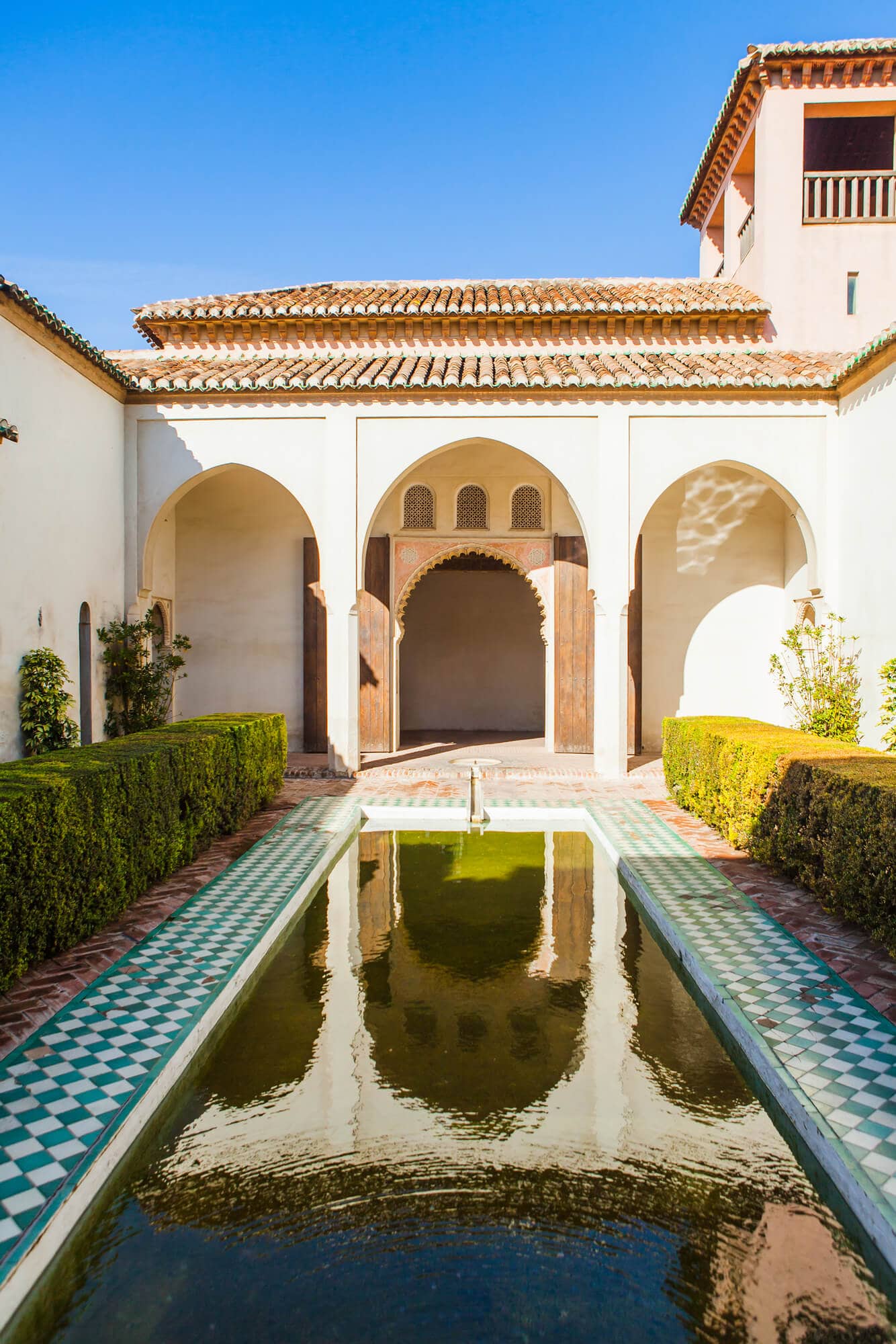 A typical Moorish courtyard set around a rectangular pool surrounded by green tiles and manicured hedges with arches mirroring in the water, inside the Alcazaba in Malaga's Old Town.