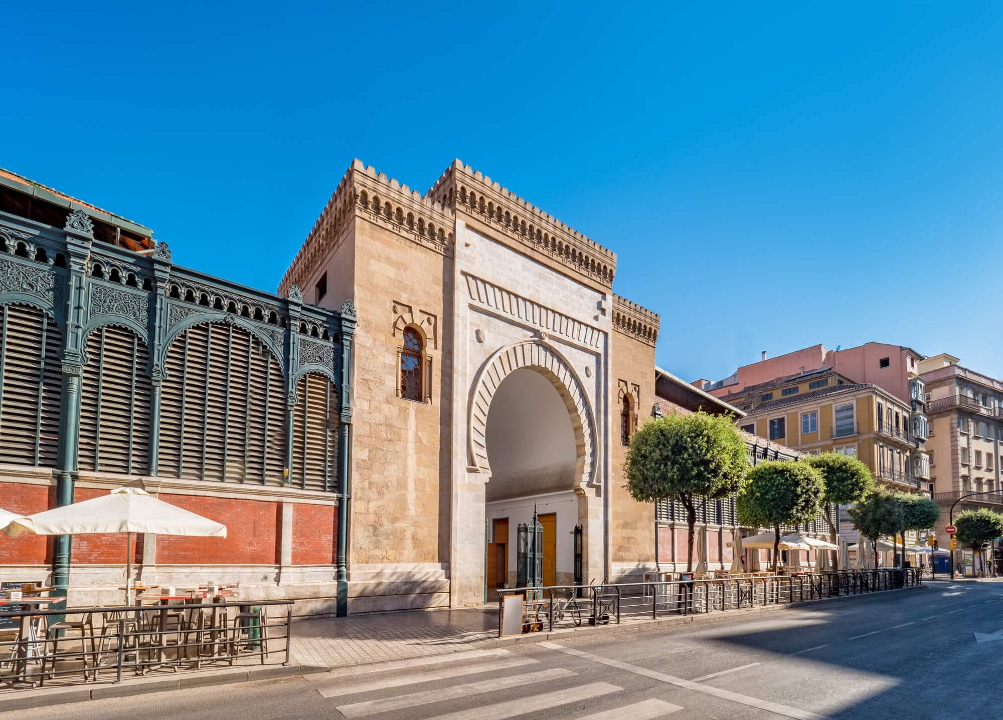 The entrance arch to Atarazanas Market in Malaga Old Toen built from sandstone in a Moorish style, with a black iron fence to both sides and manicured round trees to the right.