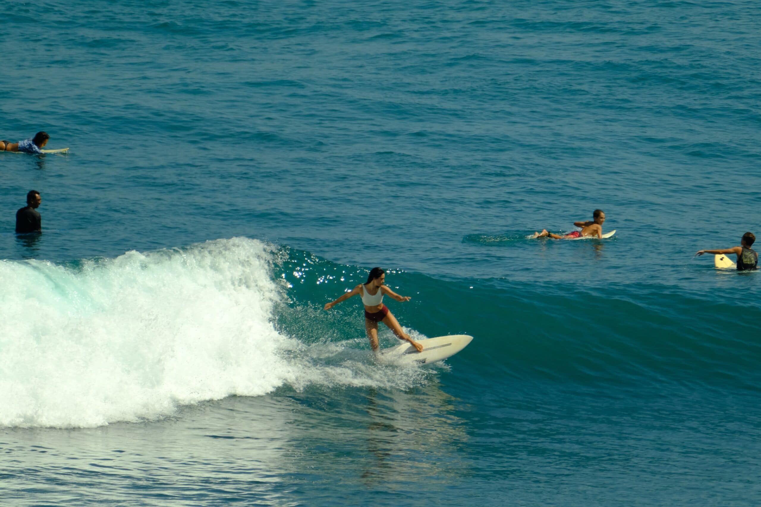 Woman surfing a wave at Echo Beach on a white surfboard, wearing a white top and red bottom, with four other surfers in the water around her.