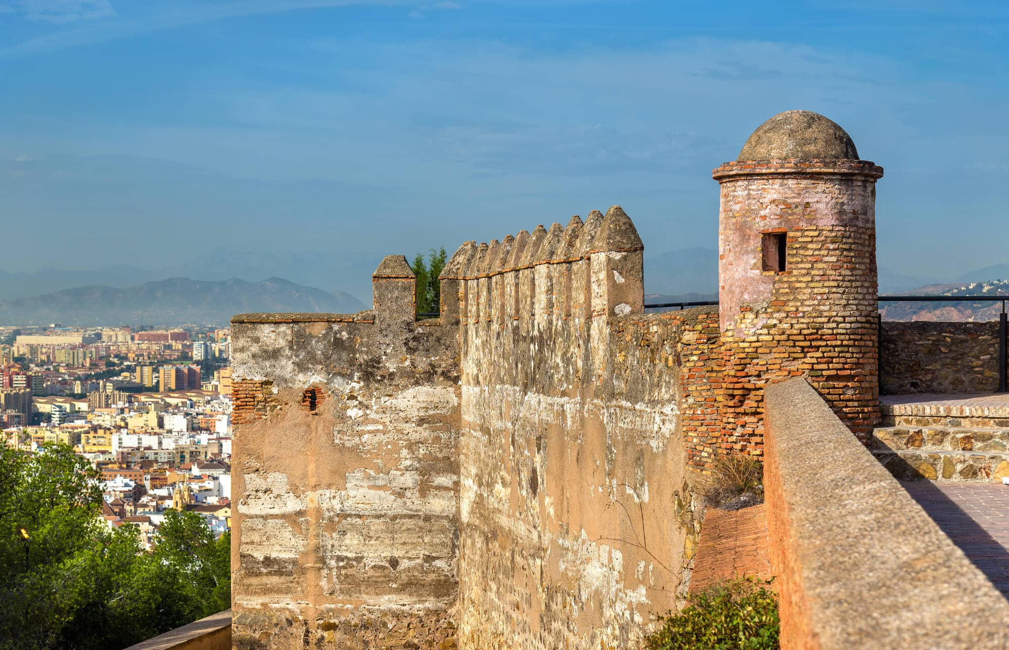 View of the outer wall of Gibralfaro Castle, one of the best viewpoints in Malaga, overlooking the city and mountains in the background with blue skies above. A must while exploring Malaga Old Town.