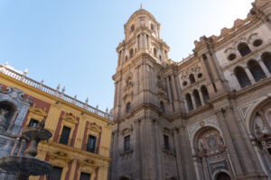 View of the ornate Malaga Cathedral from below looking up at the one tower, with the red and yellow Bishop's Palace to the left.