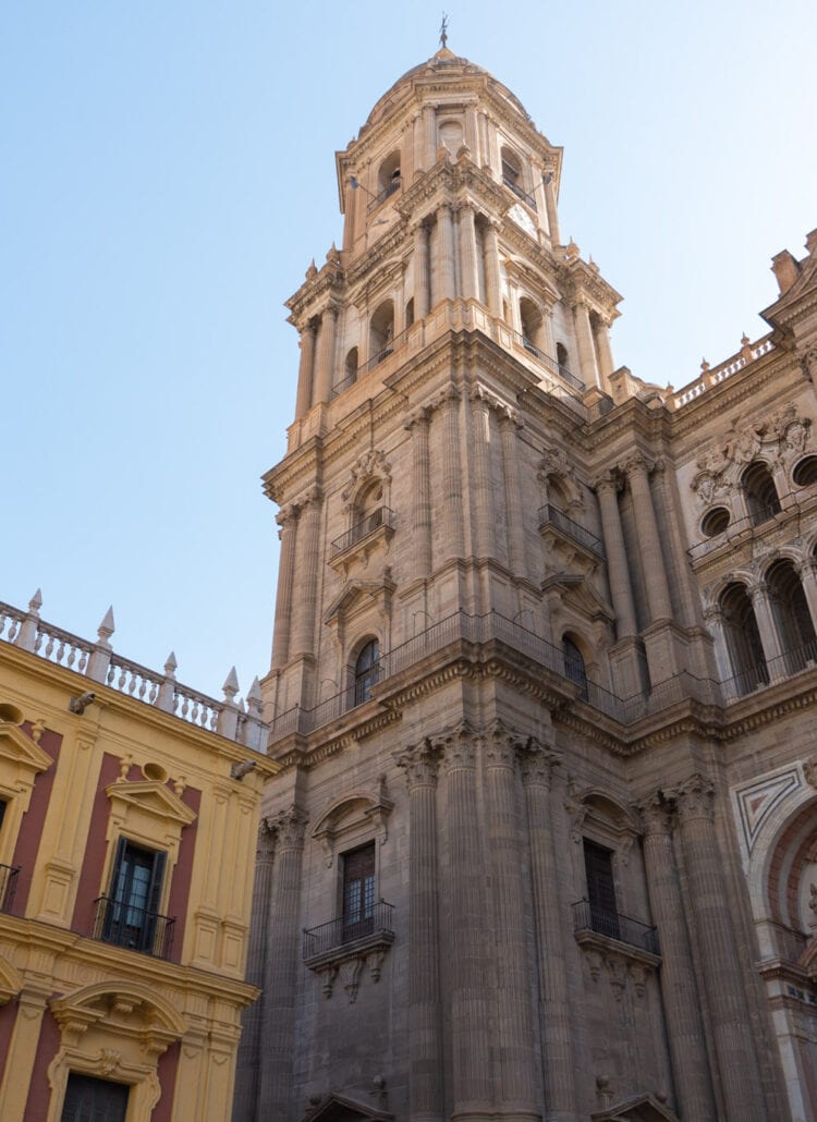 View of the ornate Malaga Cathedral from below looking up at the one tower, with the red and yellow Bishop's Palace to the left.