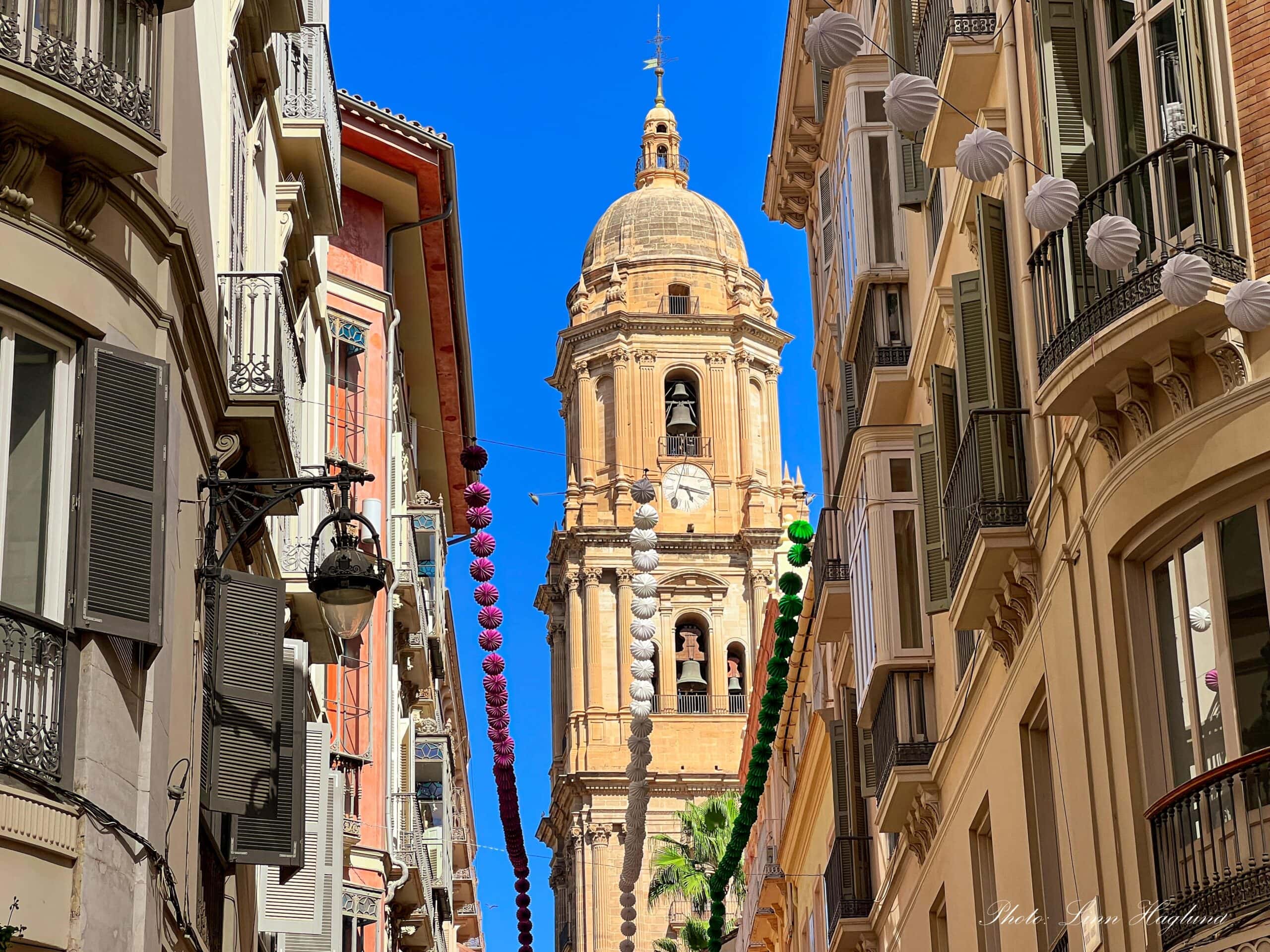 View of the one tower of Malaga Cathedral from a narrow alley with ornate building on either side, with a clear blue sky above.