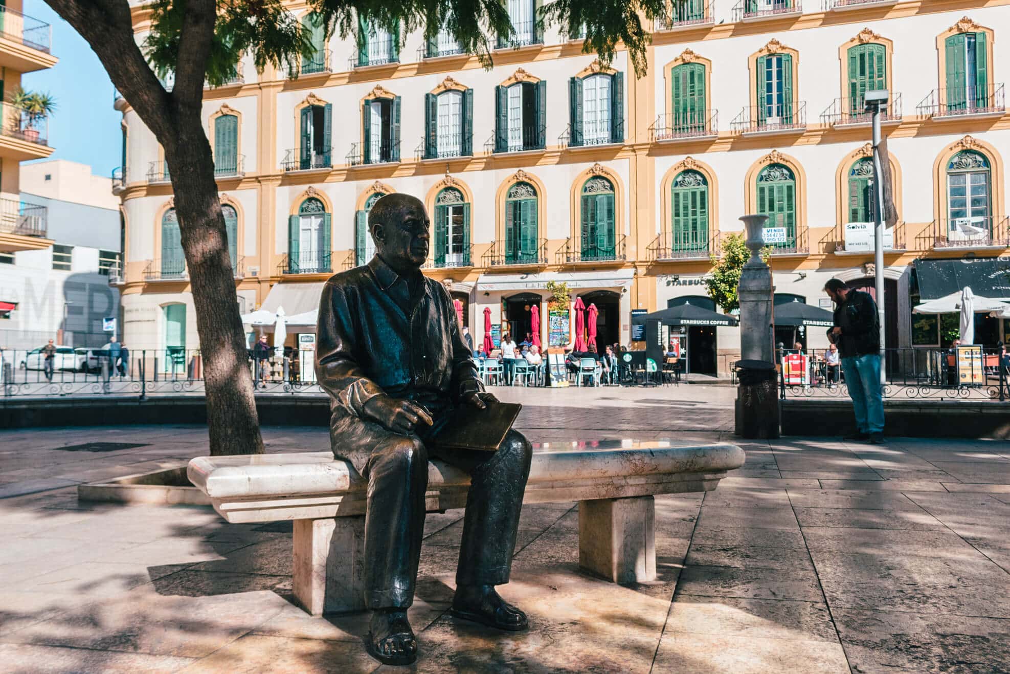 Bronze statue of Picasso sitting on a bench under a tree on a square in front of a white building with yellow trim and green windows where he was born, in Malaga's Old Town.