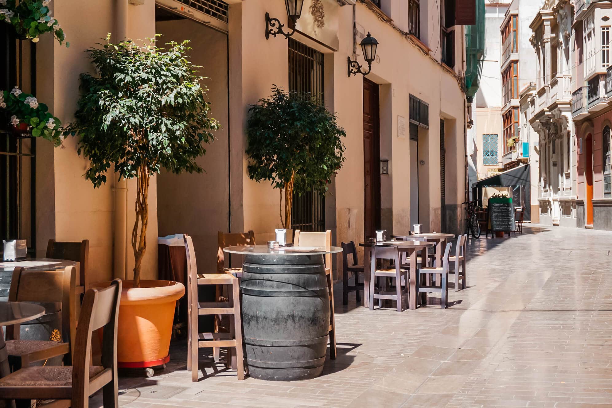 A typical taverna in a beige buidling with a few tables and chairs, an old barrel and two potted trees outside on the pedestrian street, in Malaga Old Town.