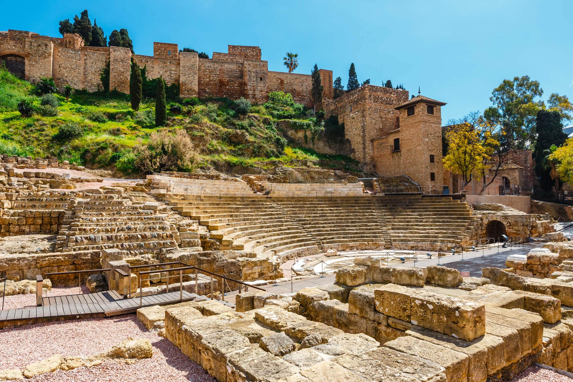 View of the old Roman Theatre ruins in Malaga Old Town with the Alcazaba and greenery in the background on a sunny day.