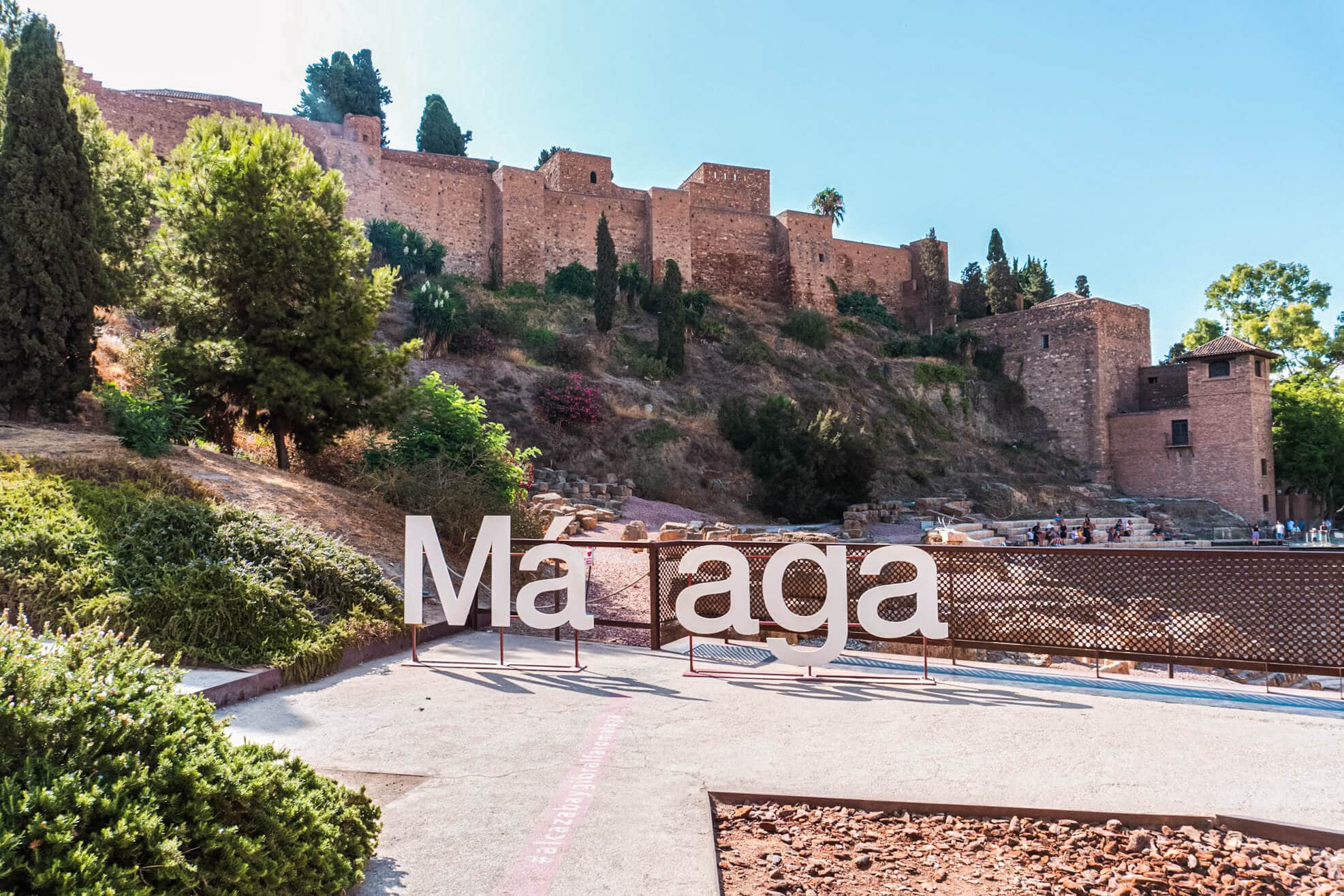 White Malaga sign with a missing l for people to stand in, in front of the Alcazaba Fortress on a sunny day.