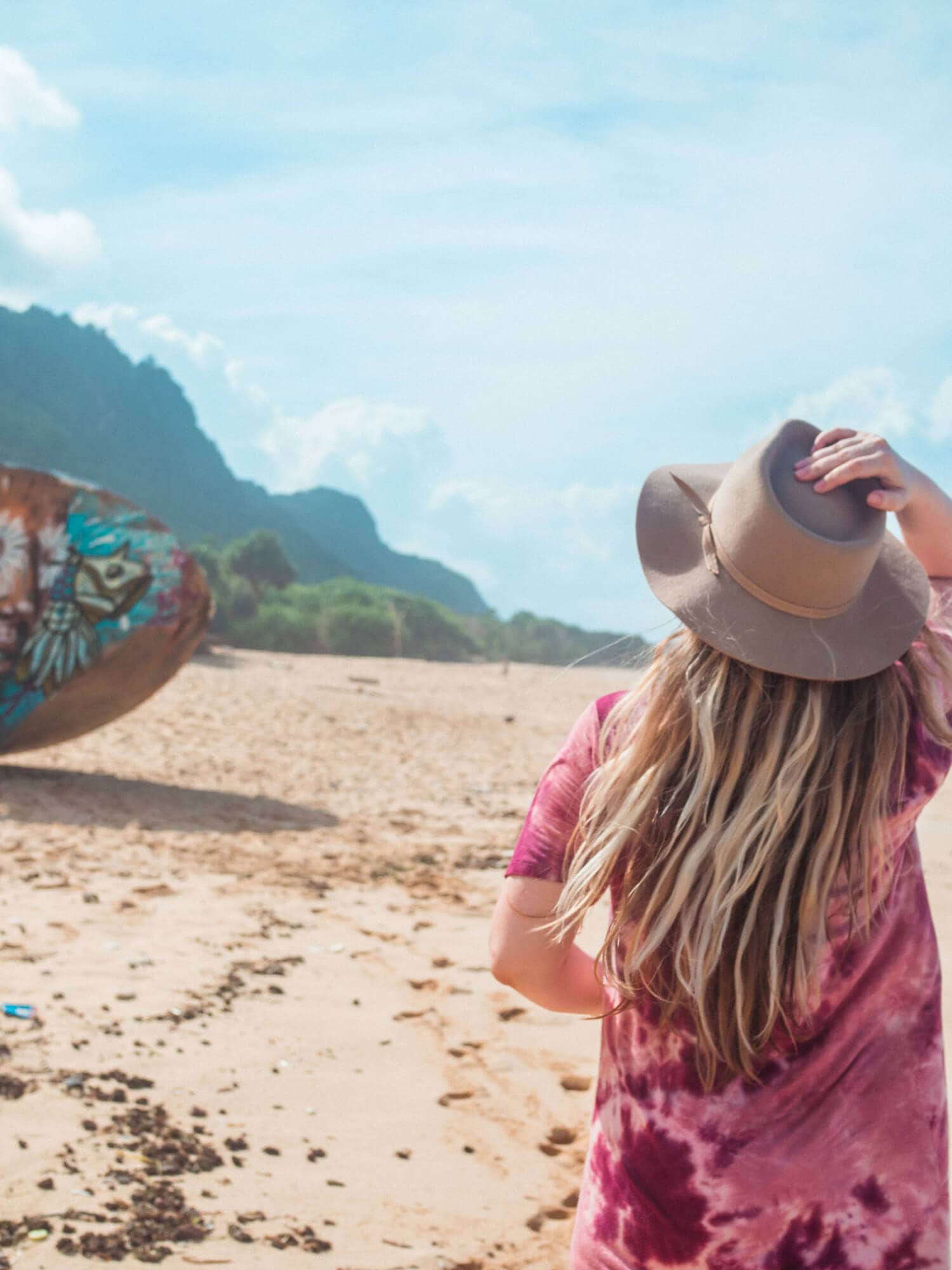 Girl with long dark blonde hair, wearing a purple tie dye dress and beige hat, walking towards a shipwreck on Nunggalan Beach during the rainy season in Bali.