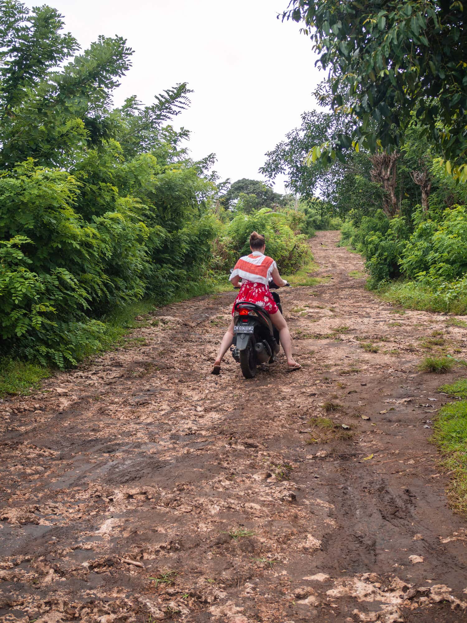 Girl wearing a short red dress and a white/orange sarong over her shoulders, driving a scooter on a steep muddy road in Nusa Penida during the rainy season in Bali.
