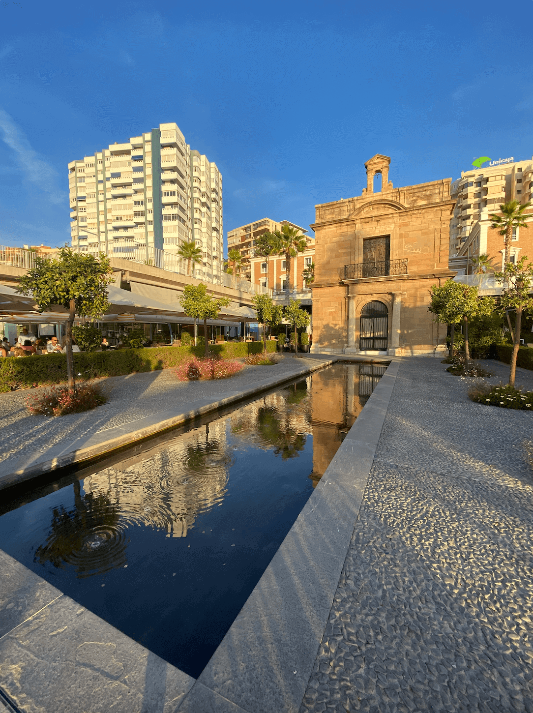A rectangular pond with an old building and a new skyscraper in the background at sunset, in Malaga's Old Town.
