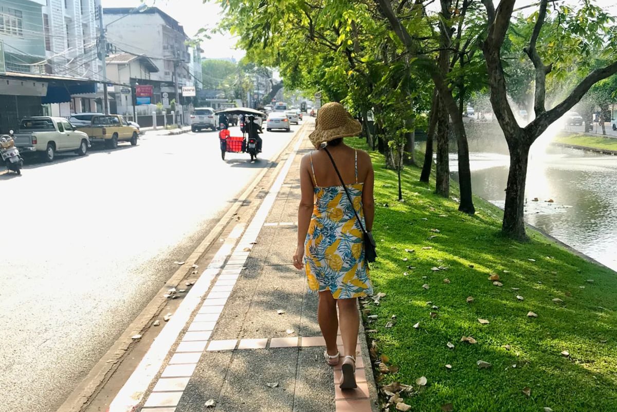 Girl wearing a brown hat, yellow and white dress and sandals walking along a road in Chiang Mai on her way to the temples.