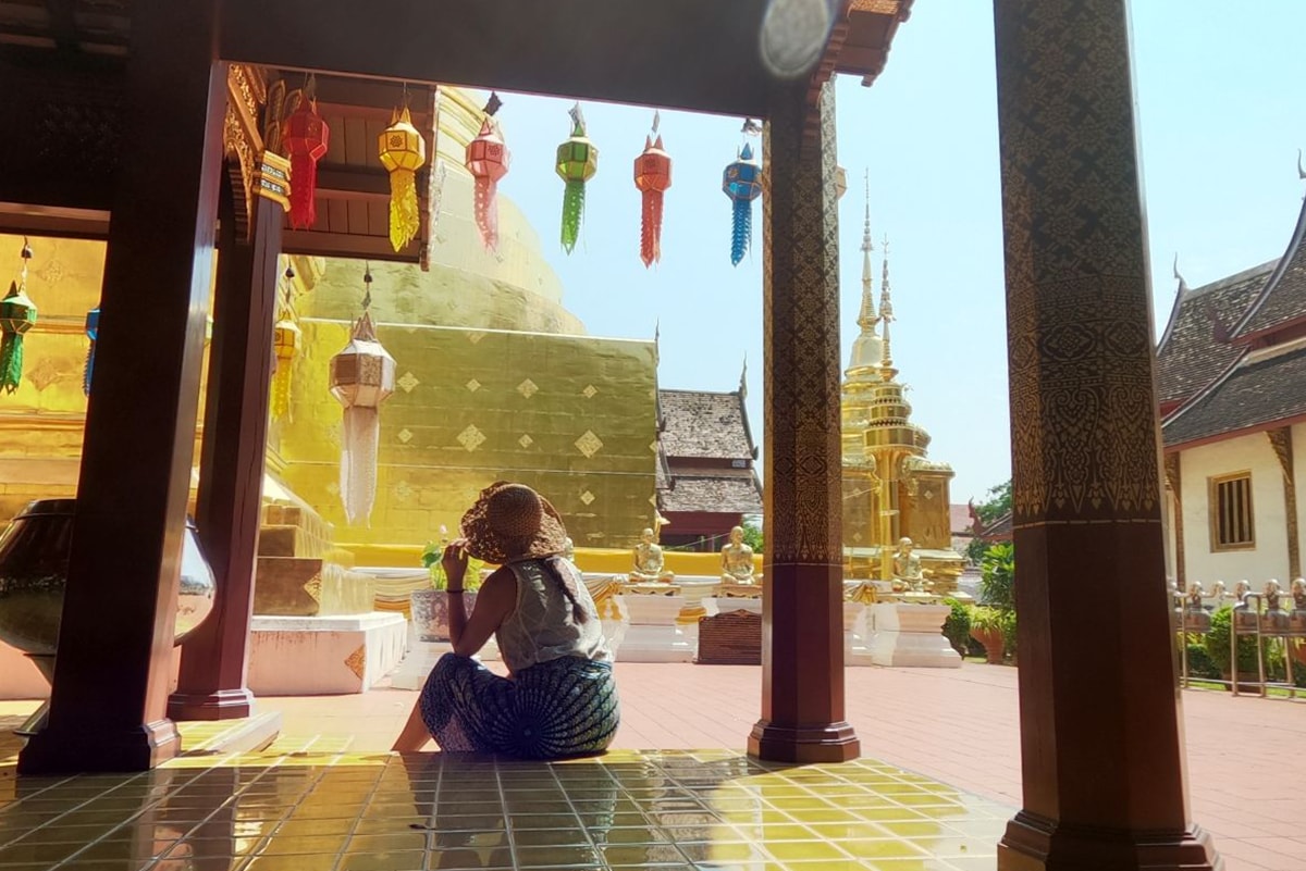 Girl wearing a hat and sarong sitting on a gold floor looking up at a large gold chedi inside Wat Phra Singh, one of the best temples in Chiang Mai, Thailand.