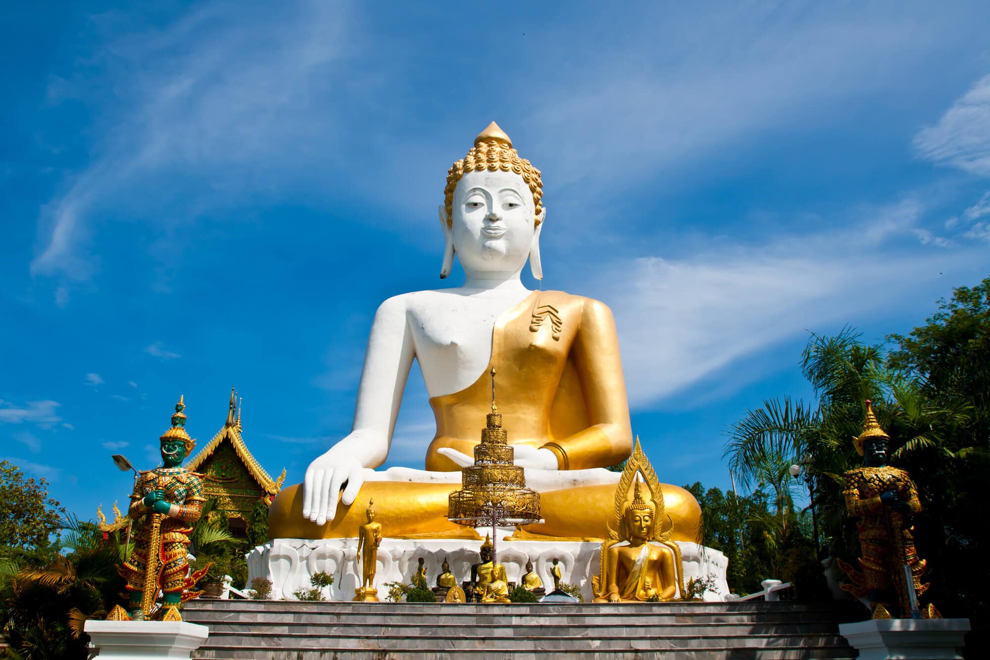 Large white and gold buddha statue with smaller statues in front set against a blue sky at Wat Phra That Doi Kham temple in Chiang Mai.