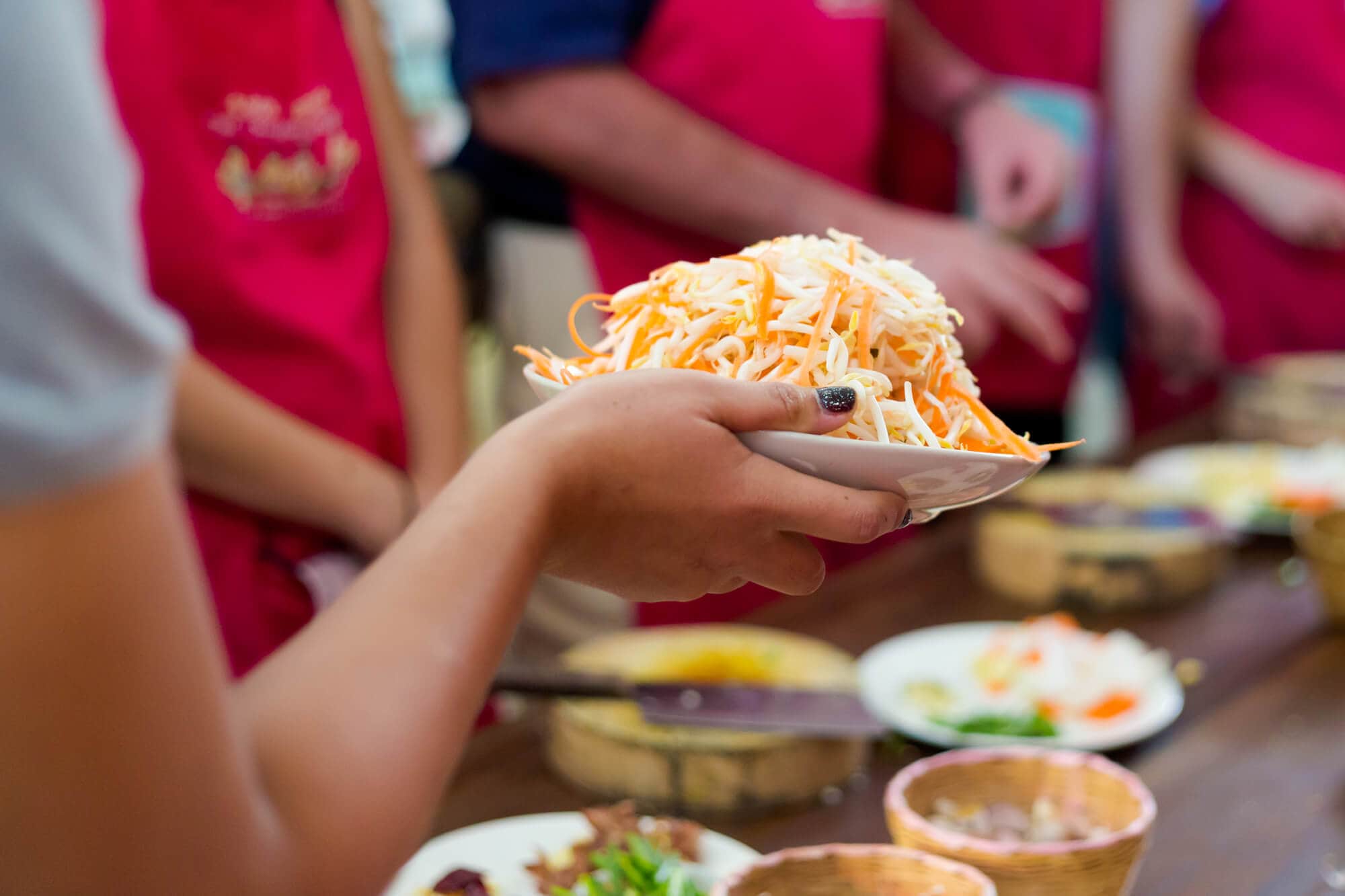 Hand holding a bowl of ingredients with people in red aprons standing in the background during a cooking class in Krabi. One of the best tour.