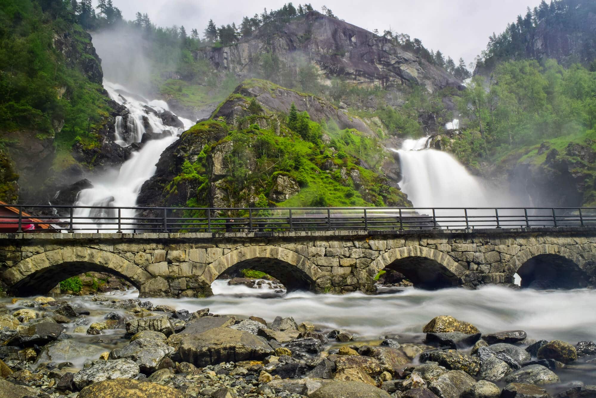 View of a stone bridge with the large Langfossen waterfall, consisting of two falls surrounded by mountains and greenery. Norway fun facts.