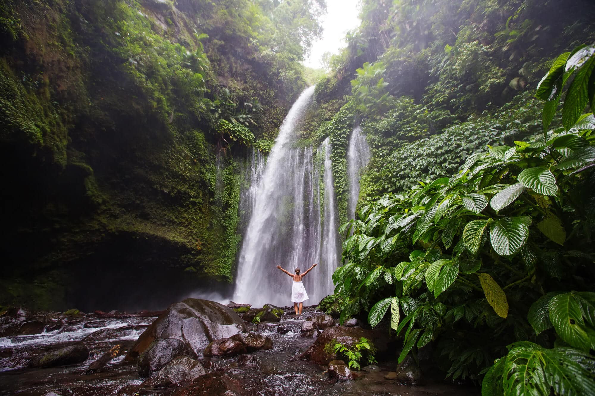 Woman in a white dress whit her arms in the air standing in front of Tiu Kelep Waterfall surrounded by lush jungle, one of the top things to do in Lombok. 