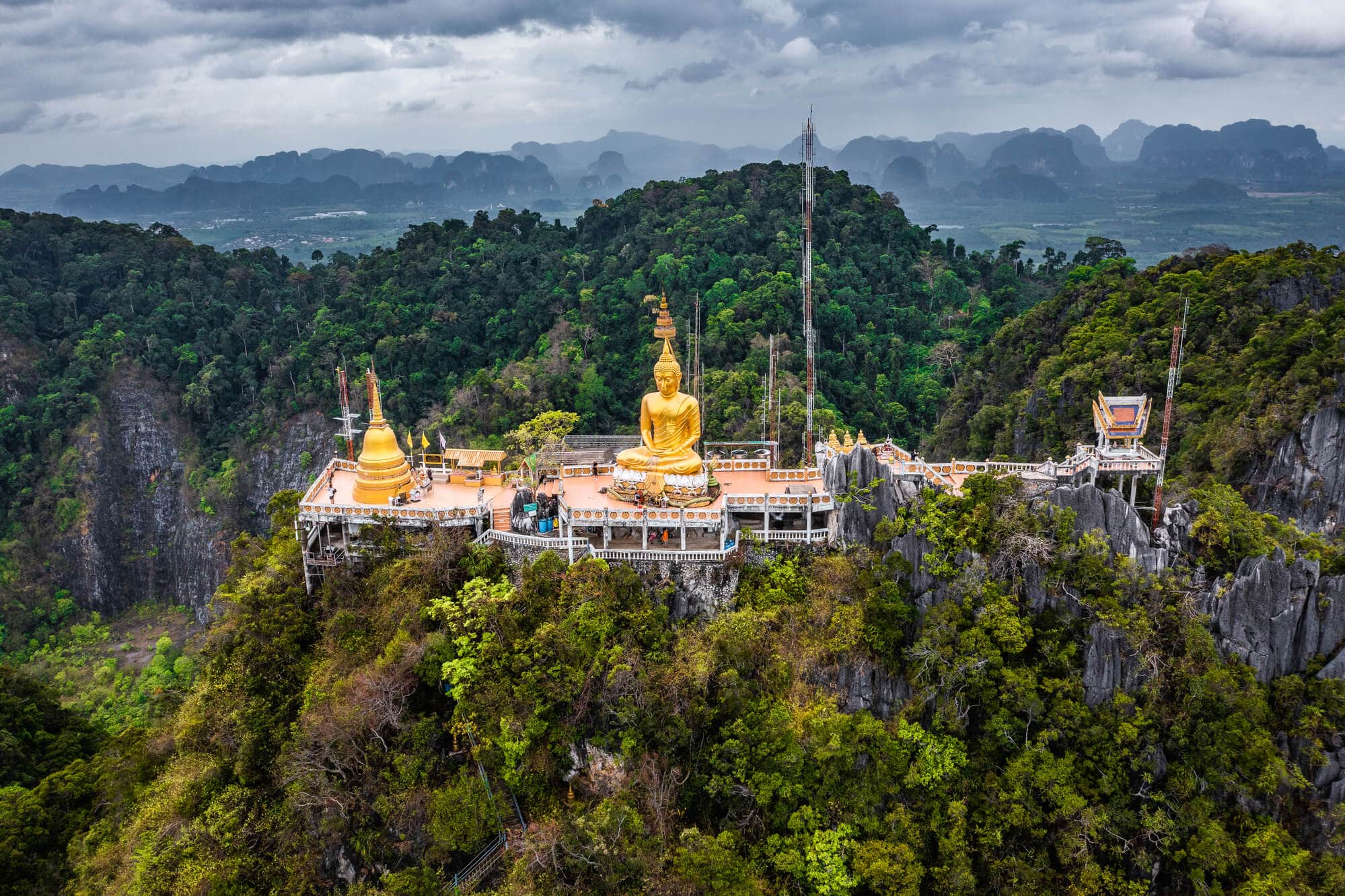 Drone view of the Tiger Cave Temple with a large gold Buddha sitting above the treet tops in the jungle with rolling hills in the background. Seen during a fantastic tour in Krabi.