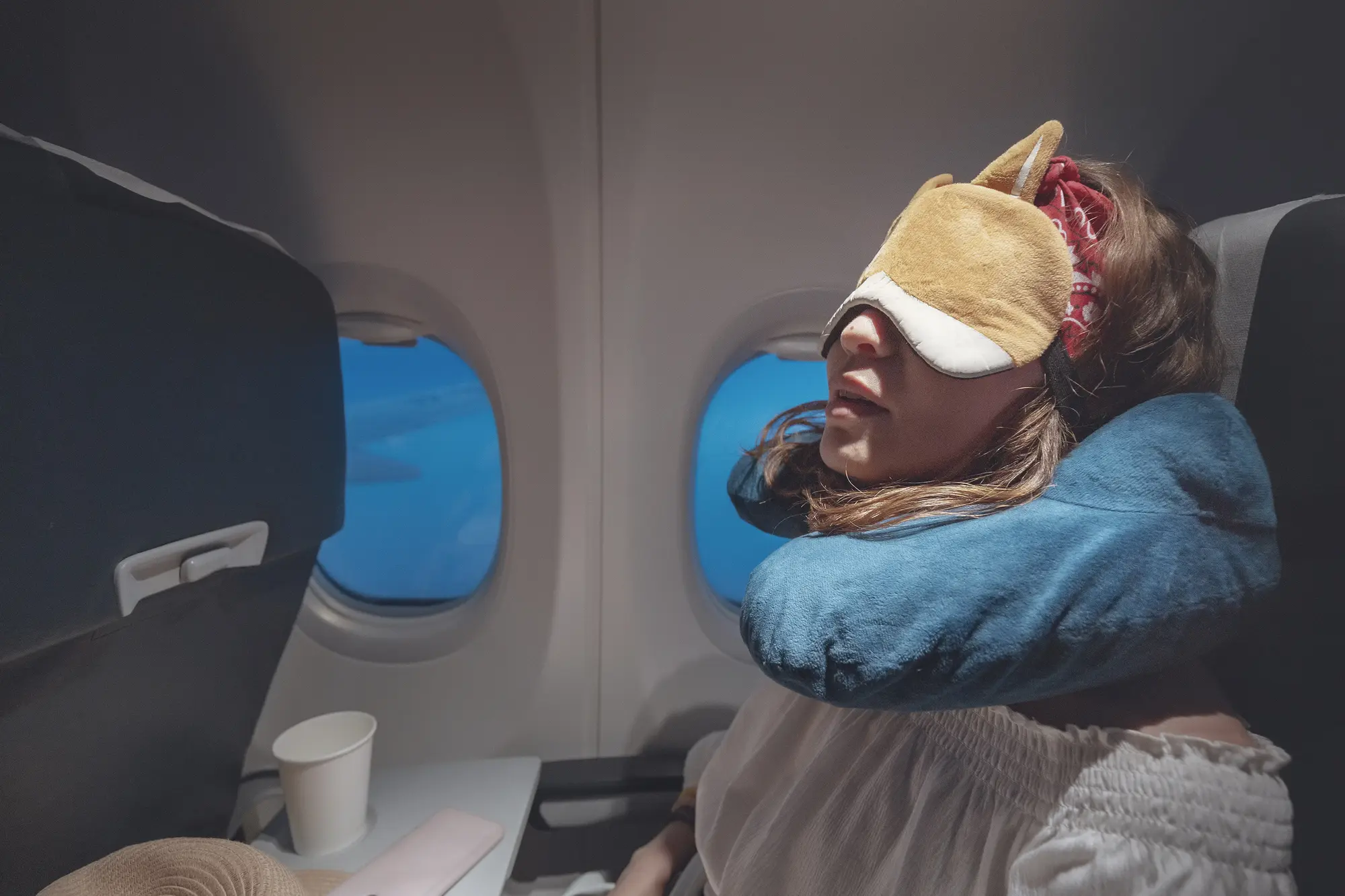 Girl sleeping on a plane in the window seat, wearing a neck pillow and sleeping mask. How to cope with fear of flying on long-haul flights.