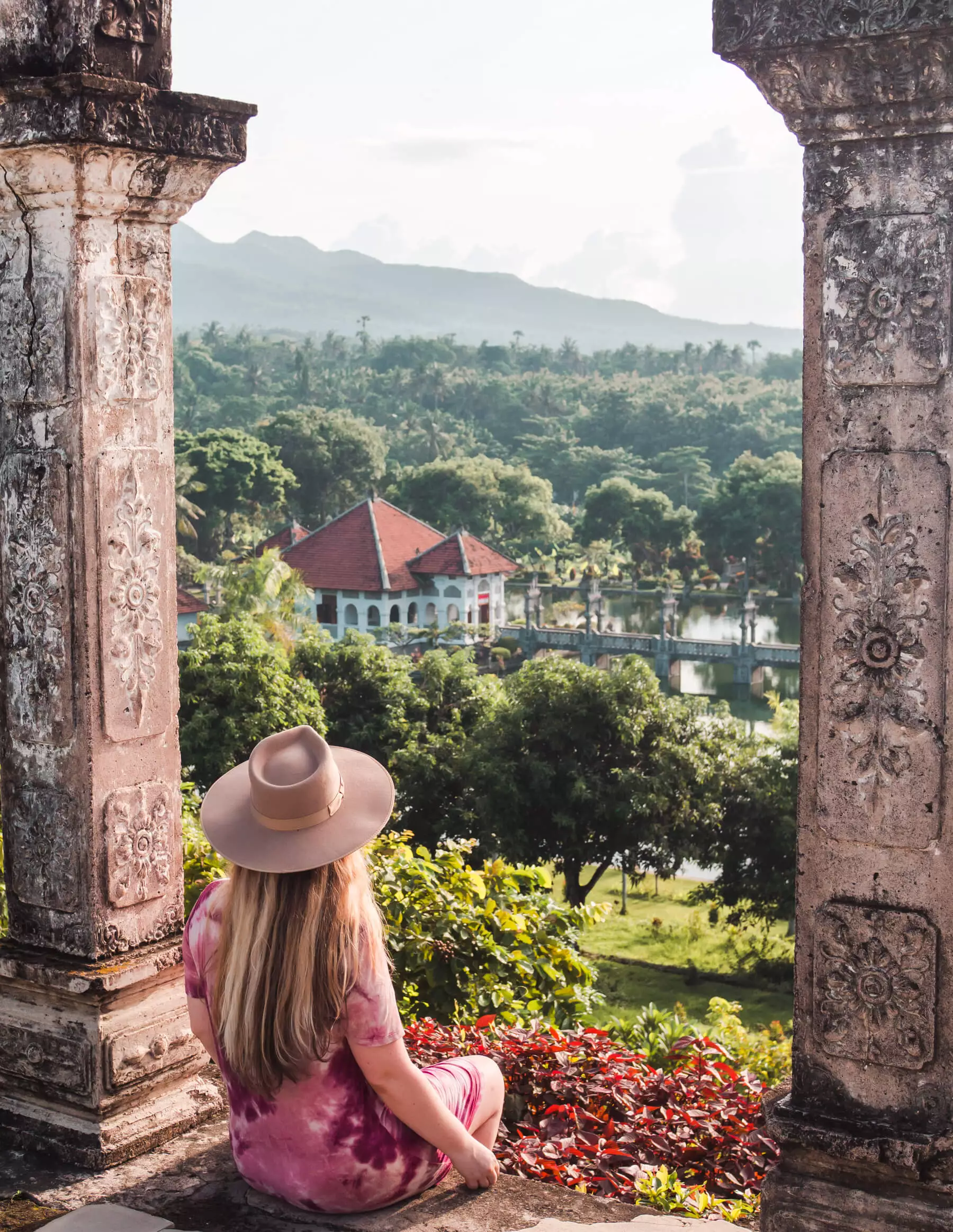 Girl in a purple dress and beige hat sitting on a platform under an arch looking out over Taman Ujung Water Palace in East Bali.