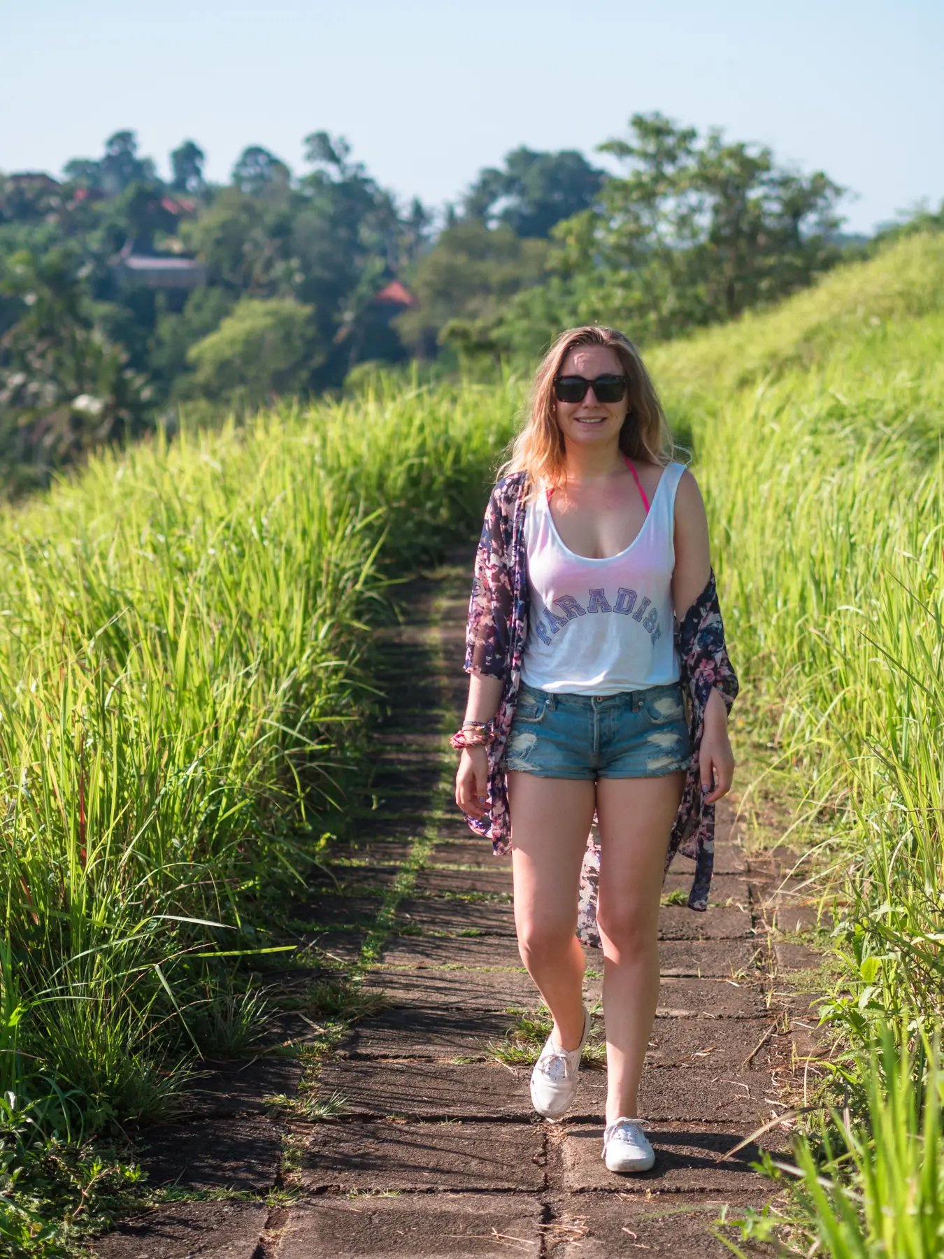 Girl wearing denim shorts, a white top and purple kimono walking along the paved Campuhan Ridge Walk in Ubud surrounded by greenery.