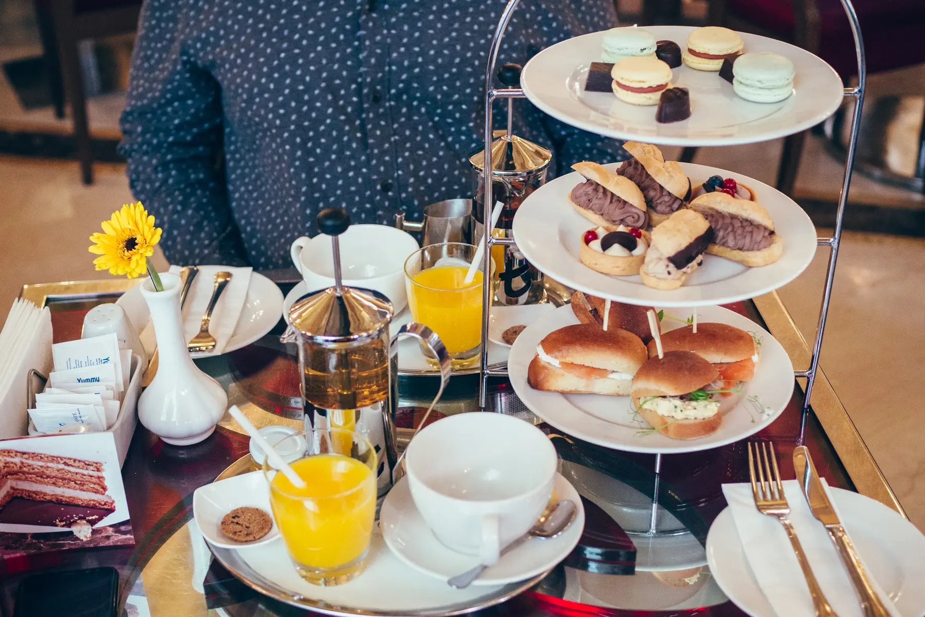 Table with a 3-tier Afternoon Tea stand with macaroons, eclairs and small sandwiches, a white plate with a glass of orange juice and white tea cup, at New York Café in Budapest.