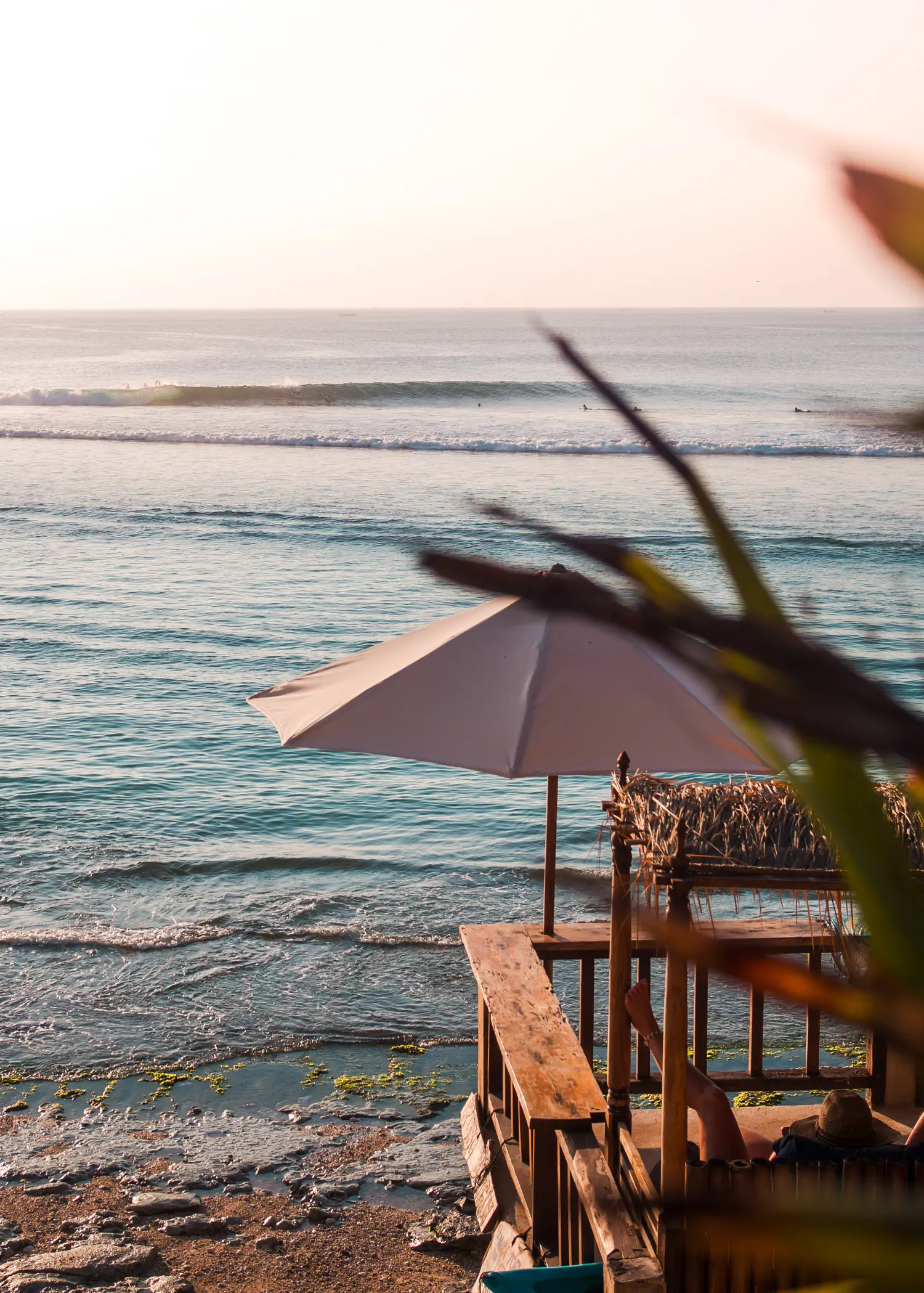 Woman sitting on a wooden veranda under a white parasol looking towards a surfer on a wave early in the morning at Bingin Beach, one of the best beaches in Uluwatu.