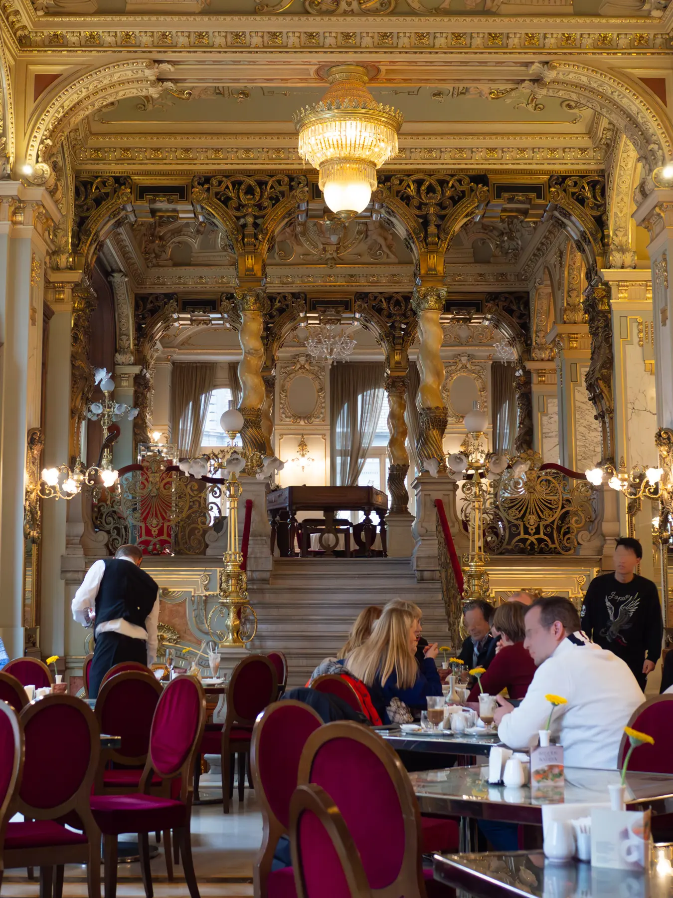 People sitting at tables on red velvet chairs inside the ornate gilded New York Café in Budapest.