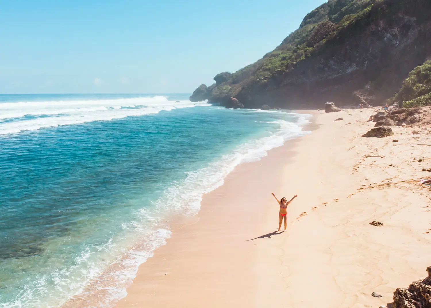 Drone view of a woman standing with her hands in the air on the deserted Nyang Nyang Beach next to turquoise ocean in Uluwatu.