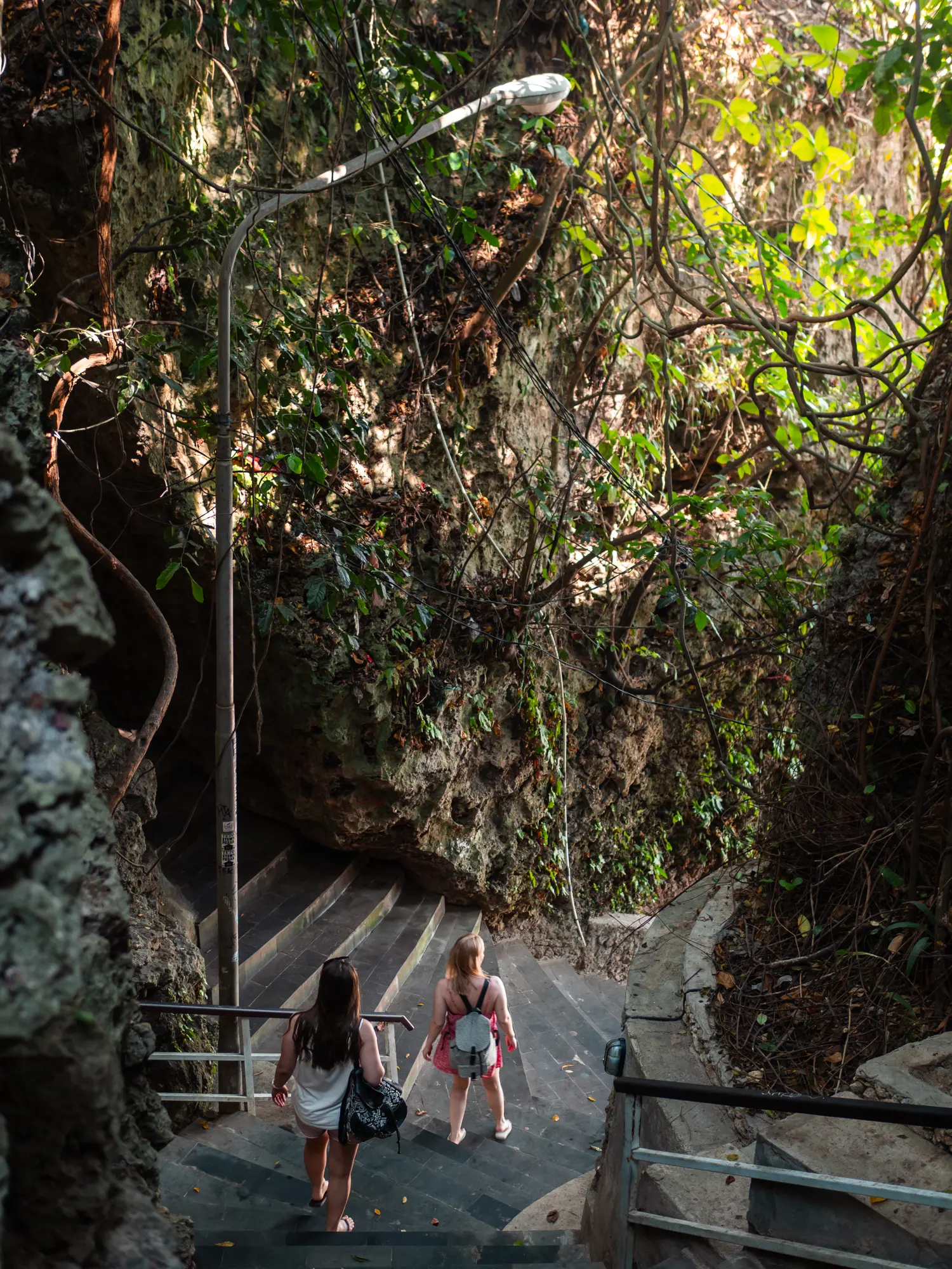 Two women wearing backpacks walking down stone stairs through the mountains at Padang Padang, one of the best beaches in Uluwatu.